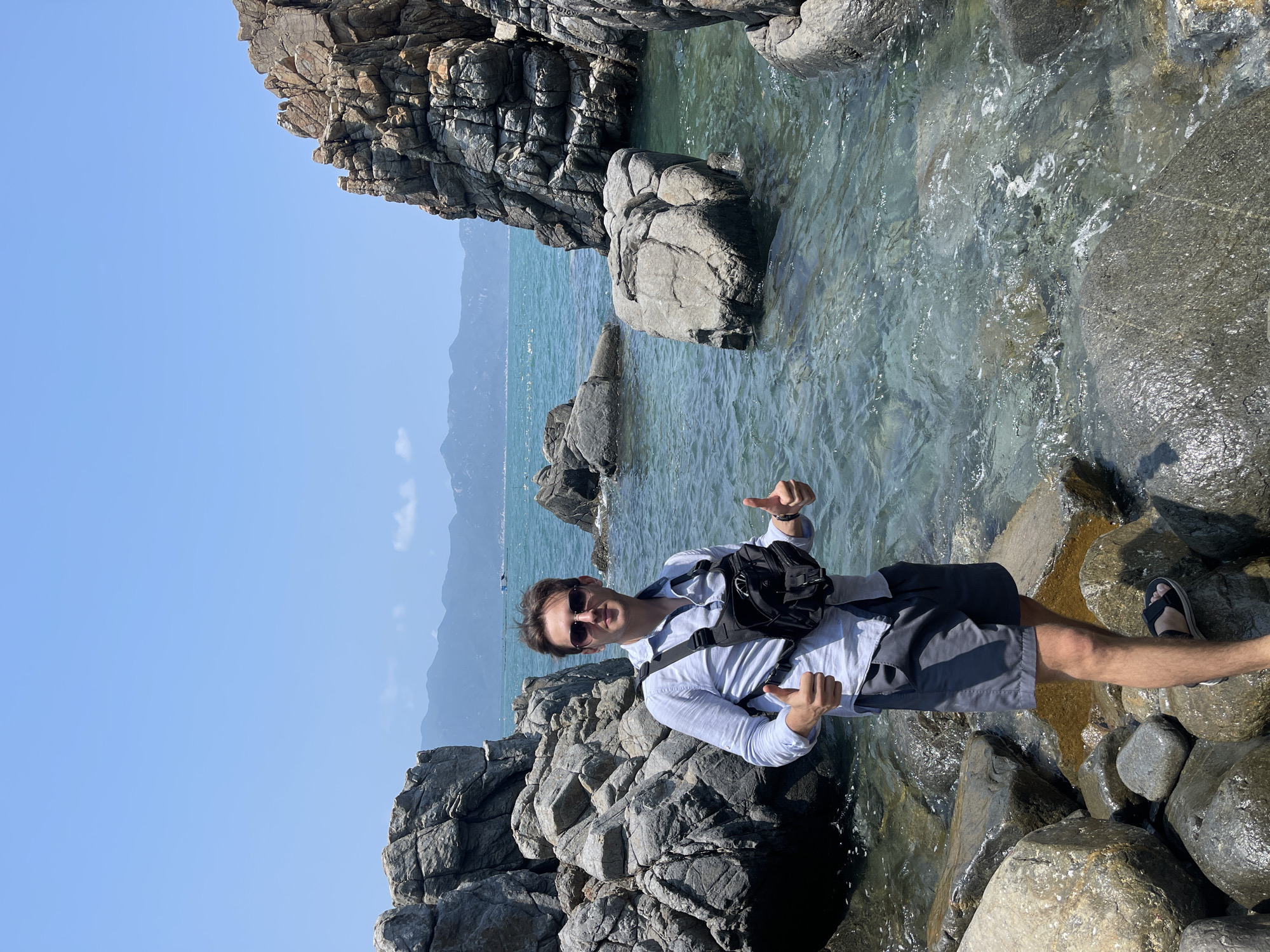 A man stands among rock formations on a beach. The water is clear and blue. In the distance are mountains. The man is wearing sunglasses, a light blue long-sleeve shirt, dark shorts, and sandals. He also has a black bag across his chest. The location is Vĩnh Lương, Vietnam.