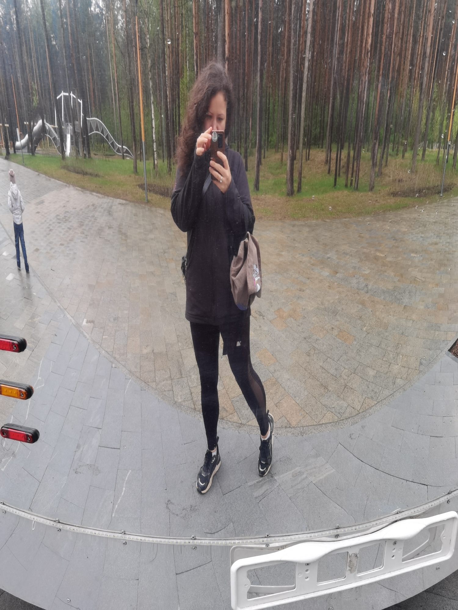 A woman is reflected in a mirror, possibly at a park. She holds a smartphone in her hands, seemingly taking a picture or using it. She is wearing a dark jacket, leggings, and sneakers, with a shoulder bag hanging on one side. In the reflection, a person walks on a path in the background, and a playground structure is visible amidst the trees. The scene is set outdoors, with a dense forest backdrop. The reflection shows a stone-paved surface and a structure with lights and a white frame at the bottom of the image. The park is a public area with playground equipment and pathways, potentially located in the city of Perm, Russia.