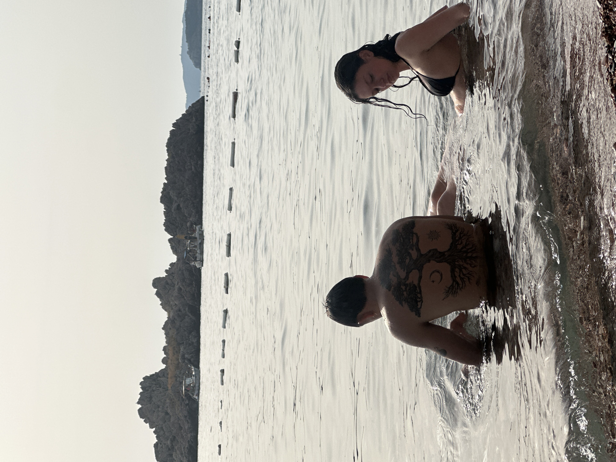 Two people are wading in the water near the shore of Sveti Stefan, Montenegro. The person on the left, facing away from the camera, has a large, intricate tattoo of a tree with a crescent moon and sun on their back. They are partially submerged in the water, with their arms resting in front of them. The person on the right, facing the camera, is wearing a black bikini top. Their hair is wet and hanging around their face. They are also partially submerged in the water. In the background, a rocky island, characteristic of Sveti Stefan, is visible, with several small boats anchored nearby. The water is calm with gentle ripples, and buoys are floating in a line across the surface. The sky is overcast, casting a soft light on the scene. The foreground shows a pebble beach.