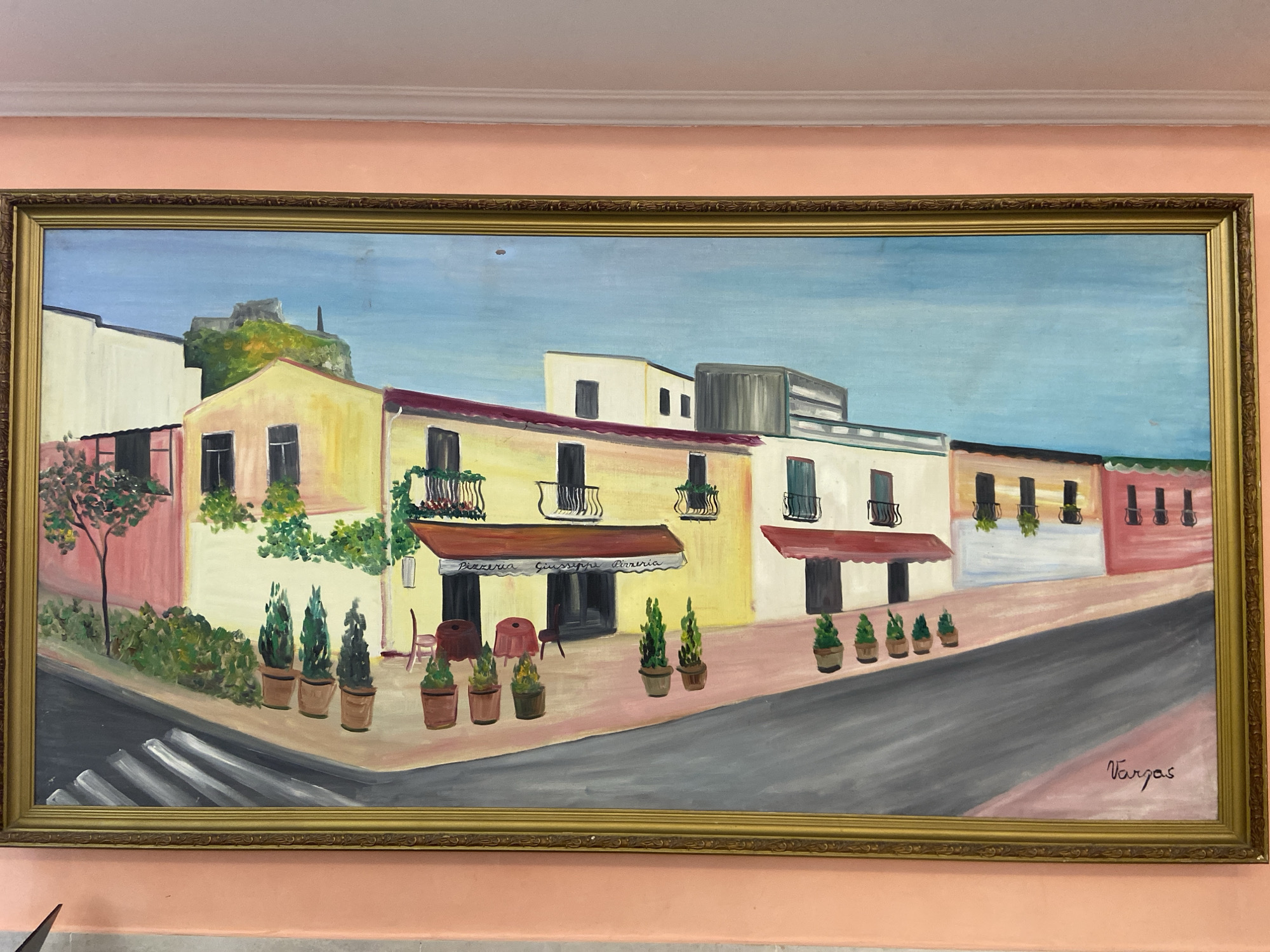 An ornately framed, horizontally-oriented painting depicts a European-style street scene. The buildings are multi-storied and feature a mix of architectural styles, painted in light colors such as cream, yellow, white, and pink. Many buildings have wrought-iron balconies and awnings over storefronts. "Pizzeria Giuseppe Pizzeria" is inscribed on one awning. Potted plants and trees are positioned along the street and sidewalk. A crosswalk is visible in the foreground, and a street slopes slightly toward the viewer. The sky is light blue. The signature "Vargas" is visible in the lower right corner. The painting is hung on a light pink wall. The location is not in Kazan, Russia.