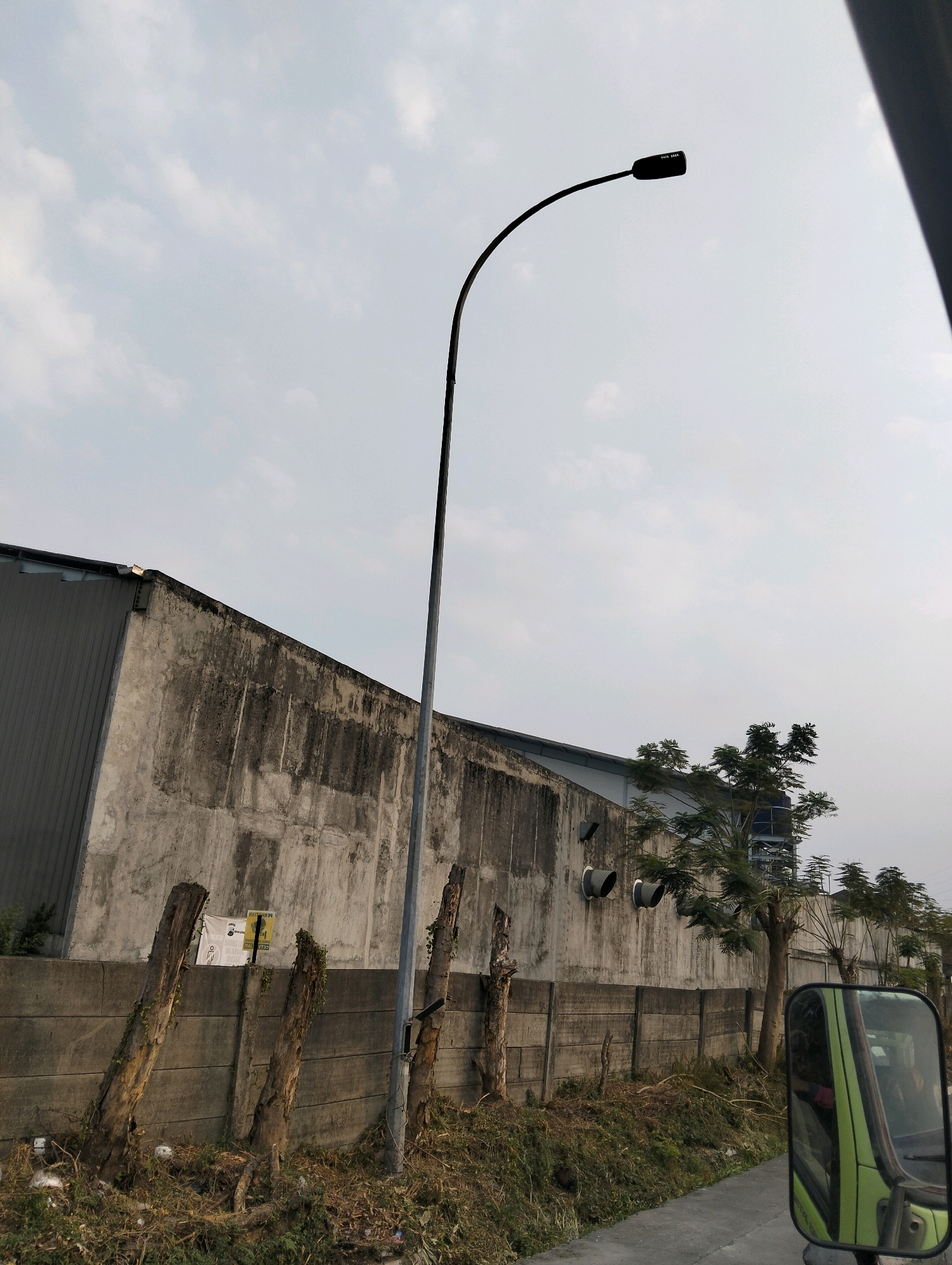 The image depicts an outdoor scene, likely on the outskirts of Semarang, Indonesia, characterized by an overcast sky. In the foreground, a light green truck mirror reflects a person's arm wearing a pink garment, suggesting the photo was taken from inside a moving vehicle. Adjacent to the road, a concrete barrier runs horizontally, separating the road from a patch of overgrown grass and dry foliage. Several weathered, upright wooden stumps are scattered along this barrier.
Behind the barrier stands a tall, weathered concrete wall of an industrial building, its surface stained with dark grime. Two circular vents are visible on this wall. To the right of the wall, a street lamp with a curved arm extends upwards against the sky. Beyond the wall, several trees with dense foliage are planted, and a water tank is partially visible behind them. The overall impression is one of an industrial or semi-industrial area with a touch of neglect.