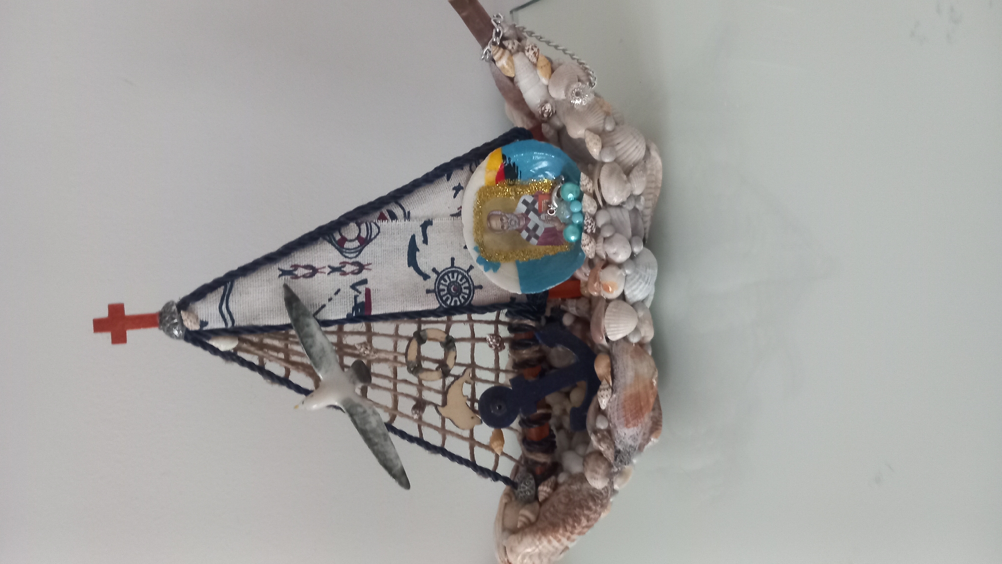 The image shows a handmade decorative boat model on a white wall. The boat is covered in seashells and has a white sail with nautical-themed patterns. A small plastic seagull is perched on the sail's netting, and a red cross is at the top of the sail. The boat's hull is also adorned with various shells. On the deck of the boat, there is a dark blue anchor, a circular decoration featuring a portrait of a man in religious attire within a blue and yellow frame, and a few blue beads. The background is a plain white wall, and the boat is positioned against it, casting a subtle shadow. The scene appears to be an indoor setting, possibly a home or a small shop. There is no specific indication of its location in Άγιος Νεκτάριος, Greece.