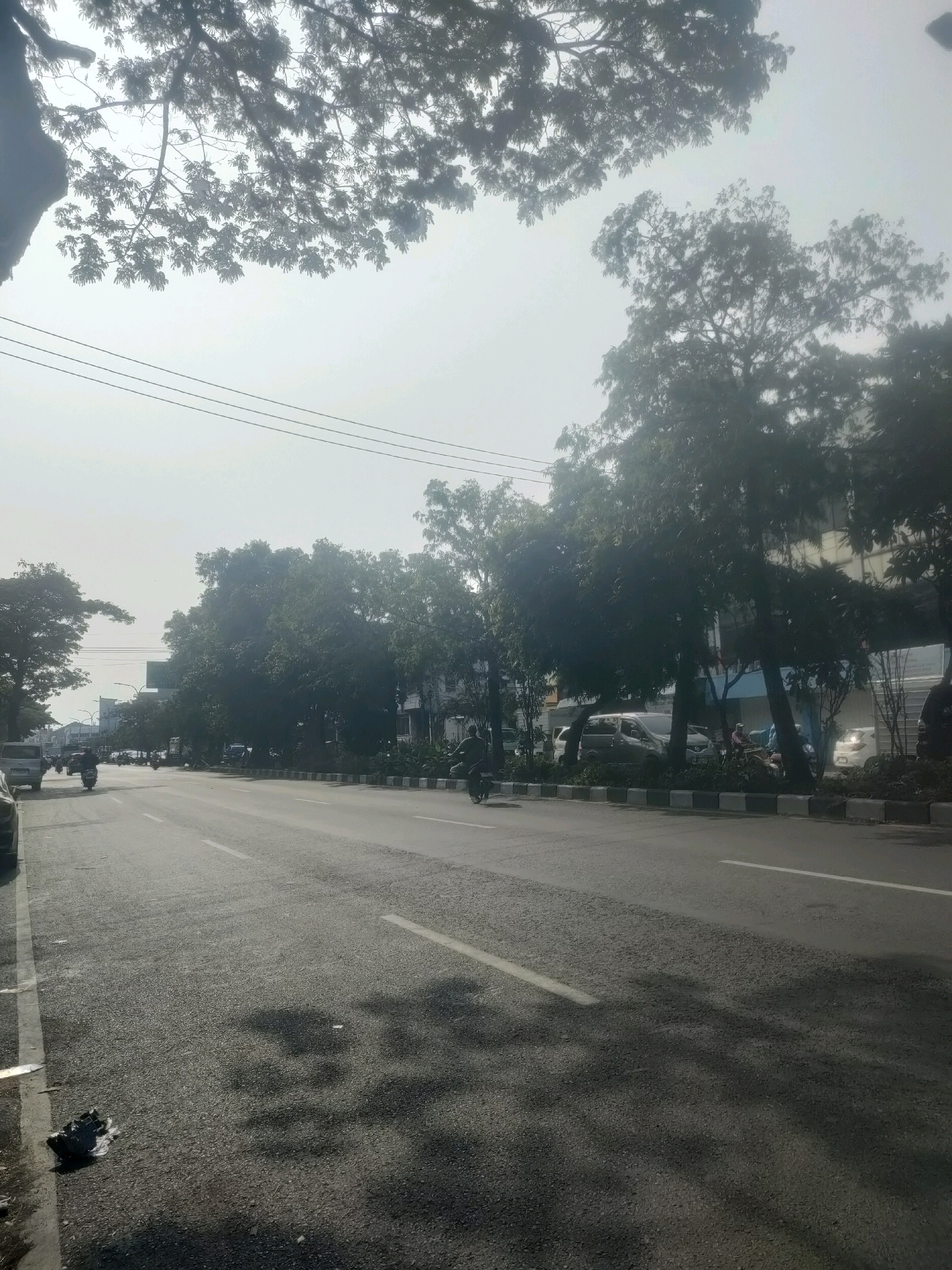 The image captures a street scene in Surabaya, Indonesia, likely during daytime. The foreground features an asphalt road with white lane markings. On the left side of the road, a white van is parked, and a dark object, possibly trash, lies on the sidewalk. Further down the road, several motorcycles and a few cars are visible, suggesting moderate traffic. Trees line the right side of the street, casting shadows on the road. Behind the trees, buildings are partially visible. Overhead, power lines stretch across the hazy sky, and the branches of large trees are silhouetted against the bright sky. The overall impression is of a typical urban environment with a mix of vehicles, vegetation, and infrastructure.