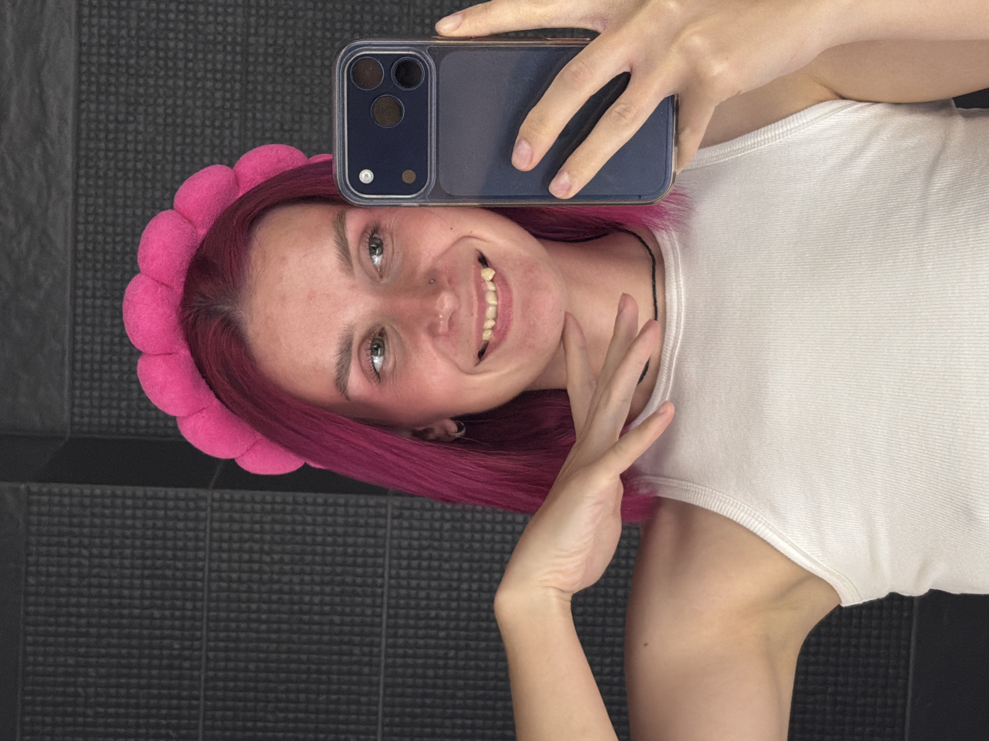 A young woman with magenta hair is taking a selfie in a bathroom. She is wearing a white tank top and a pink, fluffy headband. Her phone, a dark blue smartphone with a triple camera lens, is held up to take the picture. The background appears to be a tiled wall. The woman is smiling and touching her chin with her free hand, conveying a playful and cheerful emotion. The lighting suggests it is daytime. There is no visible text in the image.
