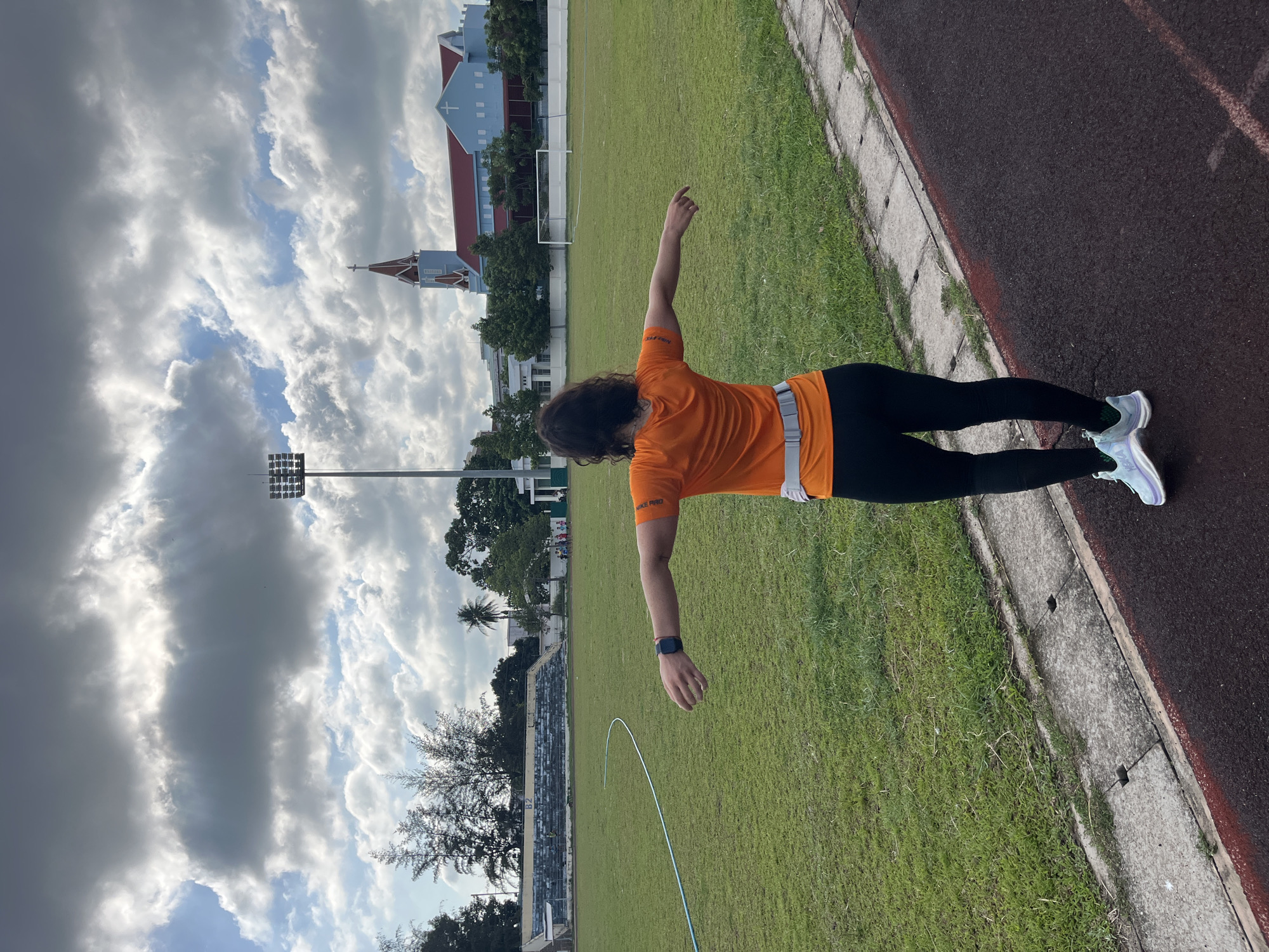 A person wearing an orange shirt, black leggings, and light blue running shoes stands on a track with their arms outstretched on a grassy field with a stadium in the background. A blue and red church with a cross on top is visible behind the stadium. The sky is filled with dramatic, cloudy formations. The scene appears to be in Phan Thiết, Vietnam, indicated by the presence of a stadium and church commonly found in the city.