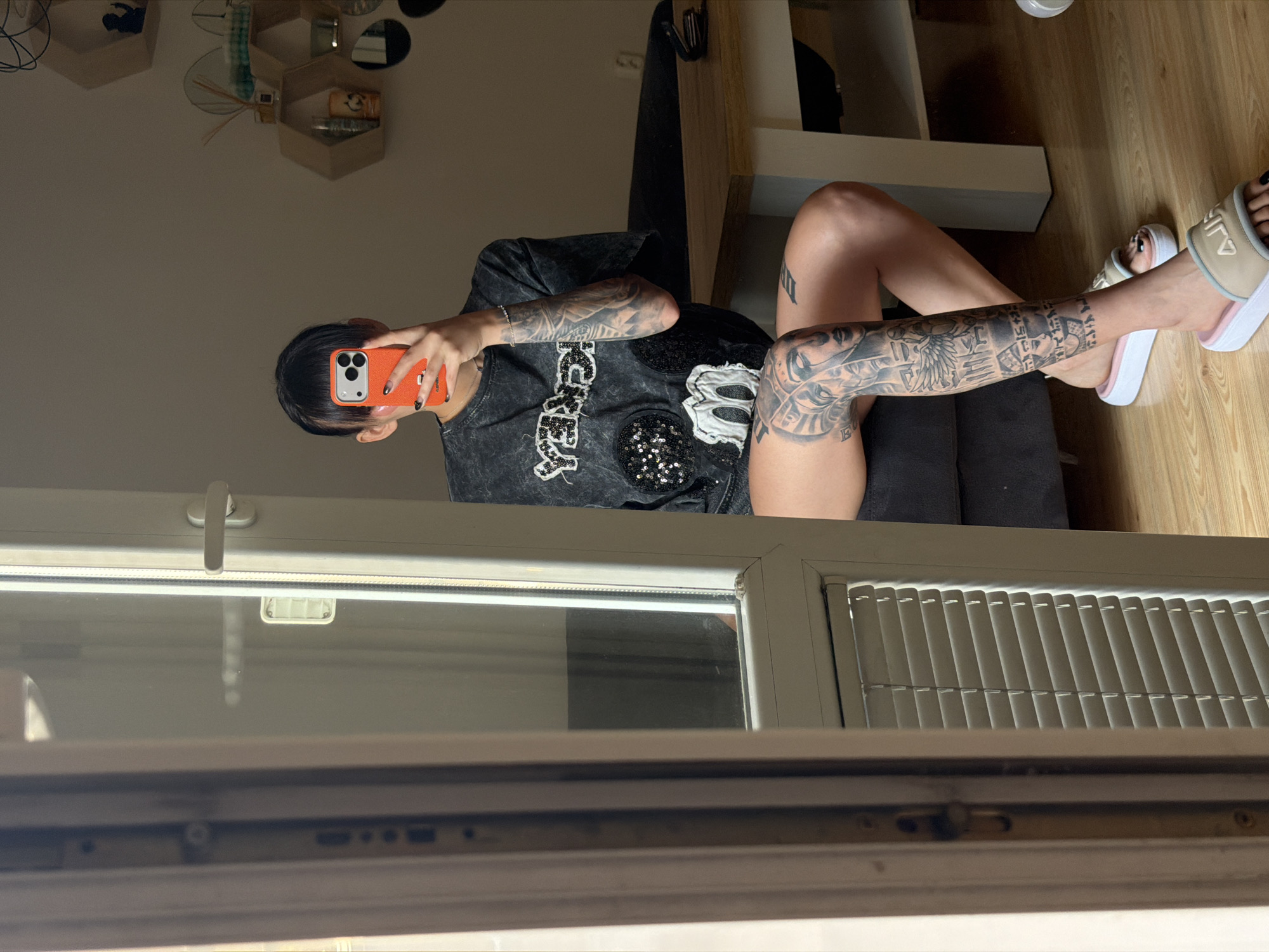 A young woman is taking a mirror selfie. She is sitting on a dark couch, with her legs crossed and extended forward. Her left leg is heavily tattooed with images that appear to be Egyptian in style, including depictions of faces and hieroglyphics. She is wearing a dark grey t-shirt with distressed fabric and black sequined embellishments, forming what looks like a character and other designs. She has short, dark hair and is holding an orange smartphone up to take the picture. Her nails are painted black. She is wearing light-colored slides with pink trim.
The woman is positioned in what appears to be a living room. A wooden table with white legs is visible to her right, and a section of a window with blinds is to her left. In the background, hexagonal shelves are mounted on the wall, displaying decorative items. The lighting suggests it is daytime, with sunlight entering the room. The location is not specified, but the scene appears to be an indoor residential setting.