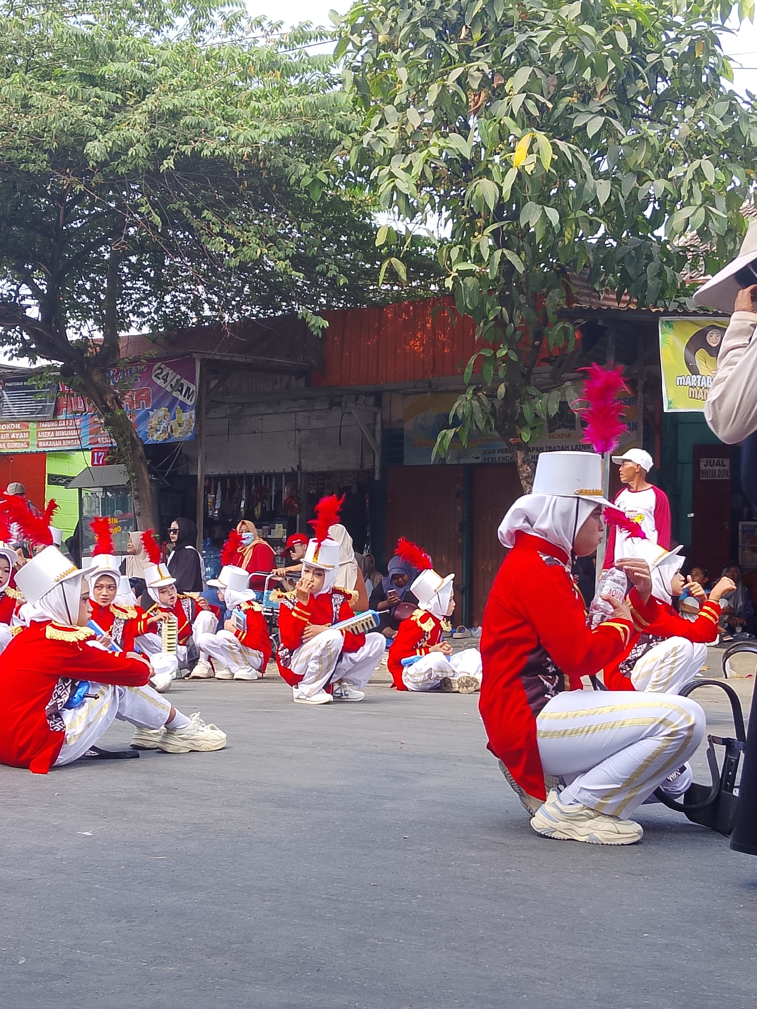In the city of Balun, Indonesia, a group of young people in marching band uniforms, predominantly red jackets with white pants and distinctive white hats adorned with pink feathers, are seen in a crouched or seated position on an asphalt surface. Their uniforms feature gold trim on the jackets and gold stripes on the pants. Some individuals are holding what appear to be musical instruments, such as glockenspiels. The backdrop consists of buildings with red and orange facades, a tree with dense green foliage, and various signs, including one with "24 JAM" and another with a yellow banner featuring an image of a person. Spectators are visible in the background, some seated and others standing, with one individual in the foreground on the right partially visible, holding a camera. The overall scene suggests an outdoor event or performance in a street or public area.