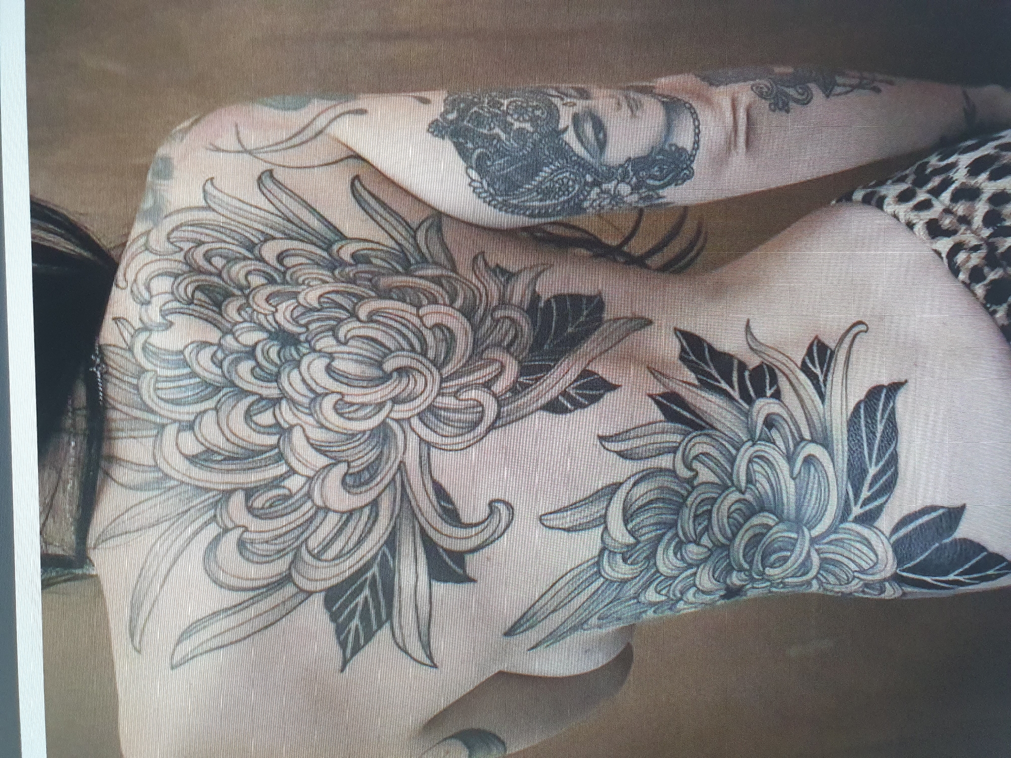 A full back tattoo of chrysanthemums is displayed on a person's back and side. The tattoo is done in black and grey ink and features intricate details of the flowers and leaves. On the right arm, a tattoo of a face with a headdress is visible. The person is wearing leopard-print bottoms. The image is taken from behind, showing the person from the shoulders down. The background is a plain, textured brown wall. There is no indication of the image being located in Tashkent, Uzbekistan or any other specific city.
