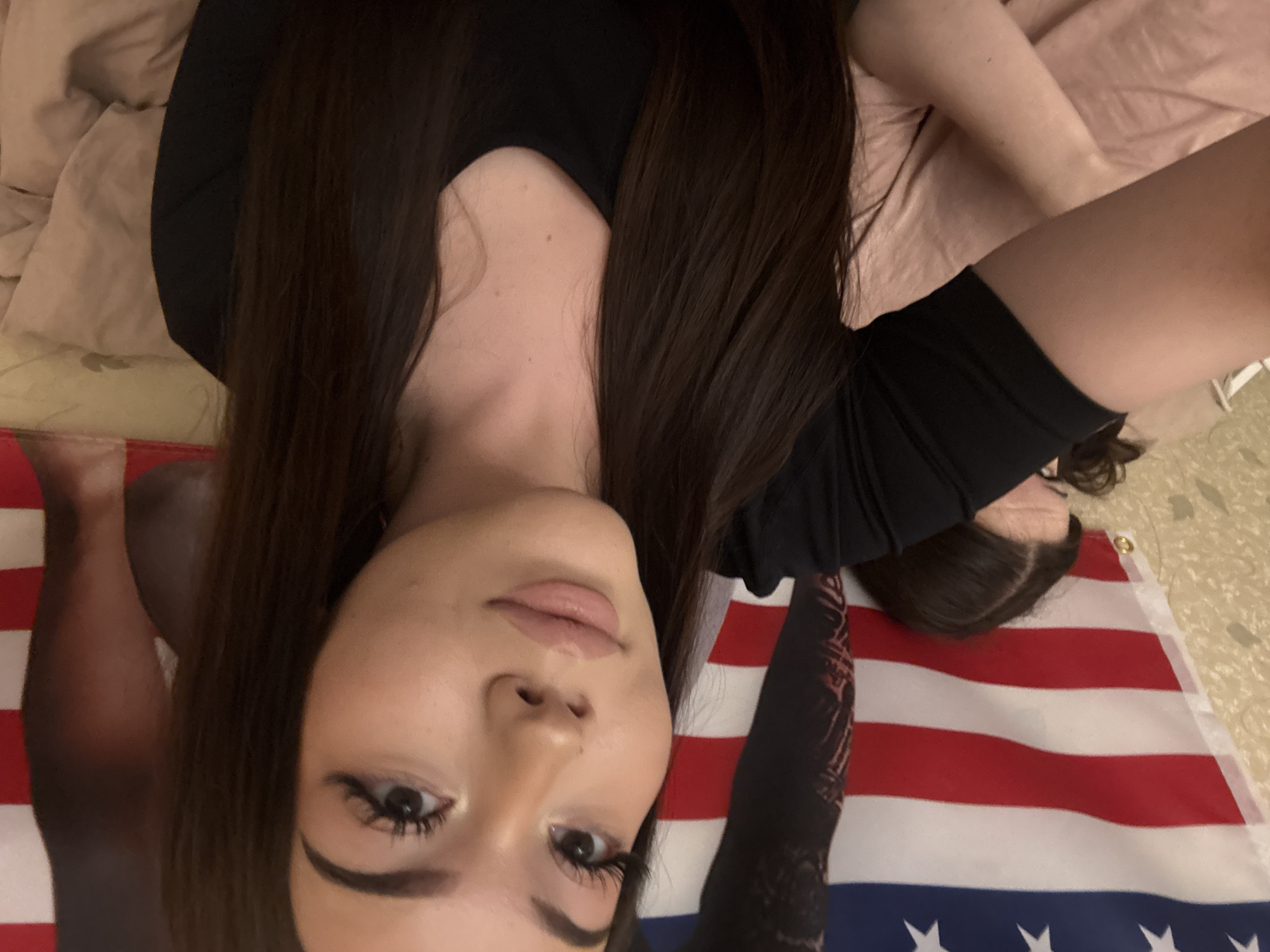 The image captures a close-up shot of a young woman lying down and looking directly at the camera. Her head is positioned towards the bottom-center of the frame, slightly angled, with her long, dark brown hair spread around her. She has a neutral expression, with prominent eye makeup including dark eyeliner and long eyelashes, and her lips are adorned with light pink gloss. She is wearing a black long-sleeved top. Her arms are visible, one extending towards the top-right of the frame, and the other partially visible on the left.
The woman appears to be lying on a light pink or beige fabric, possibly bedding. Beneath her body, a large United States flag is spread out, displaying its characteristic red and white stripes and a blue canton with white stars. The background also reveals parts of a light-colored floor or carpet. The setting is indoors, likely a room. No discernible text is visible.
