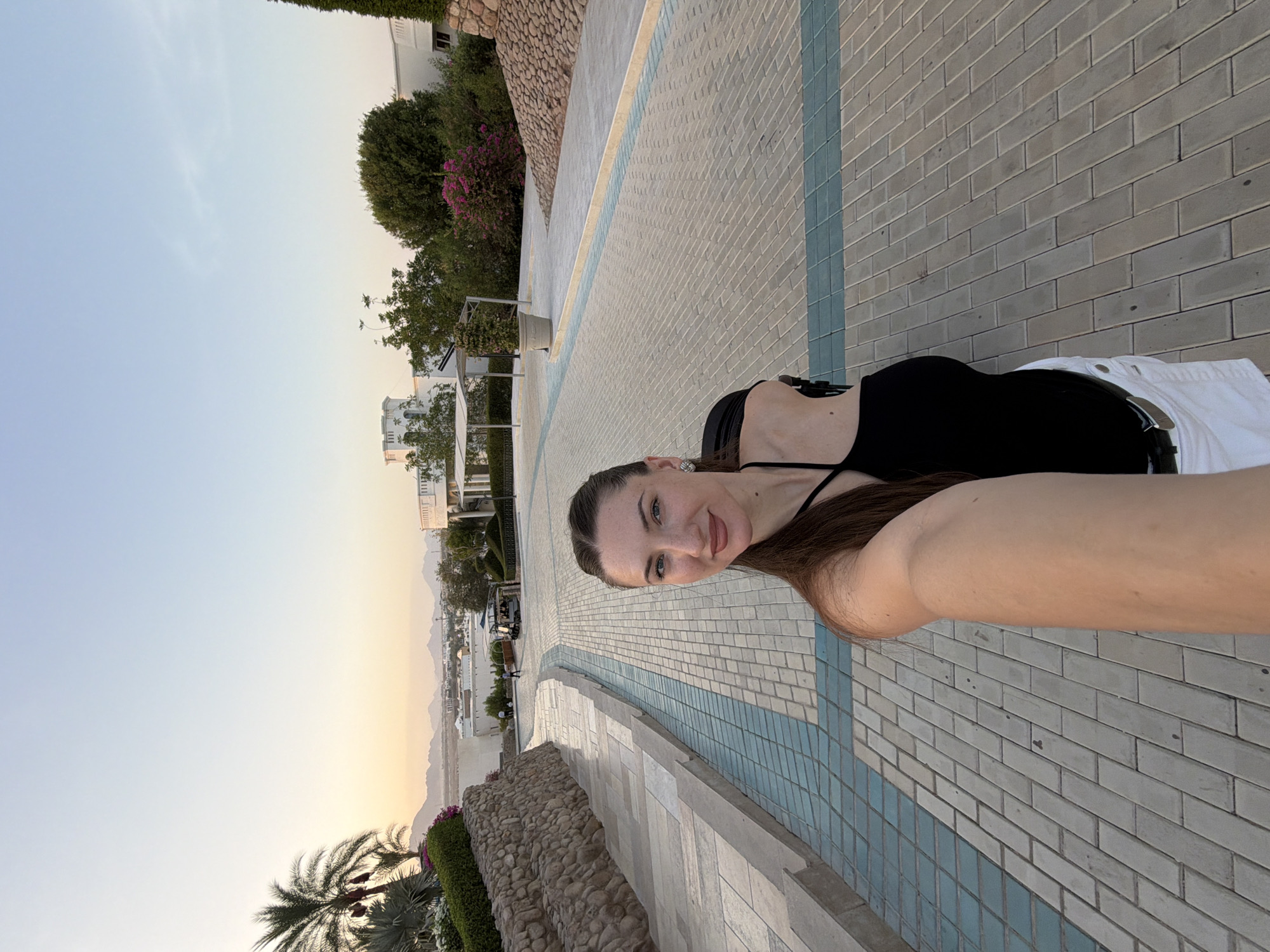 A woman takes a selfie while walking on a paved walkway in Sharm El Sheikh, Egypt, during sunset. The walkway is made of light-colored pavers with a decorative blue tile accent. To her left, a stone wall borders a sloped area with greenery and palm trees. In the background, a resort complex with white buildings and lush landscaping is visible, with mountains in the distance. The sky is a soft gradient of orange and blue, indicating the end of the day. The woman is wearing a black camisole and white shorts, and her hair is pulled back.