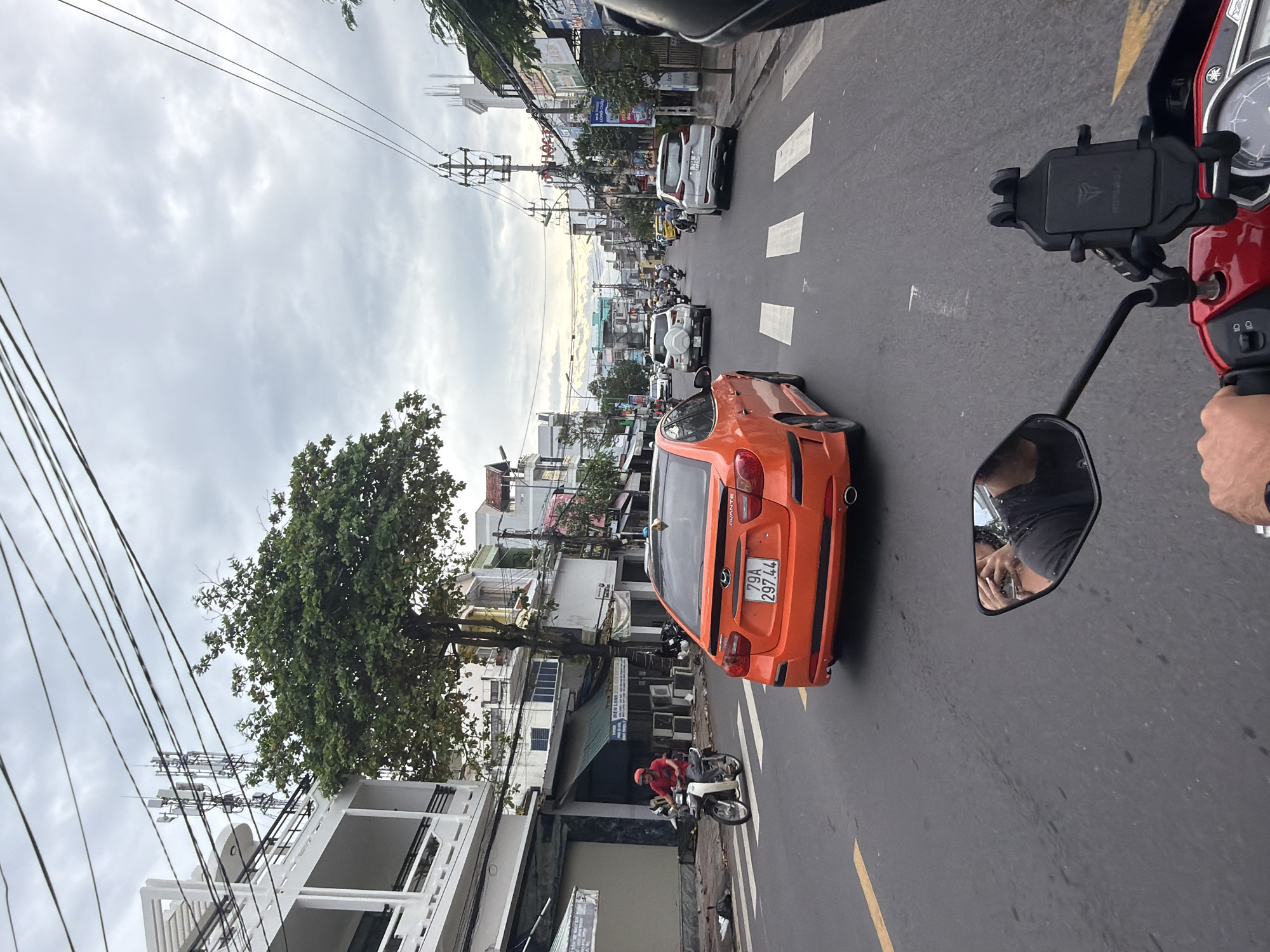 An overcast day in Nha Trang, Vietnam, captures a street scene with moderate traffic. The perspective is from a motorcycle, looking forward. In the foreground, the motorcycle's red handlebars and a black phone mount are visible. To the left, a side mirror reflects a person taking a photo. A red Hyundai Avante sedan is in the middle ground, driving on the right side of the road, with a license plate reading "79A 297.44". A man on a scooter, wearing a red shirt and helmet, rides in the left lane. Buildings line both sides of the street, with shops and residential structures. Electrical wires crisscross overhead, supported by utility poles. Several other vehicles, including cars and motorcycles, are on the road. The street has pedestrian crosswalks and lane markings. The sky is heavily clouded, suggesting an impending rain or a generally gray day.
