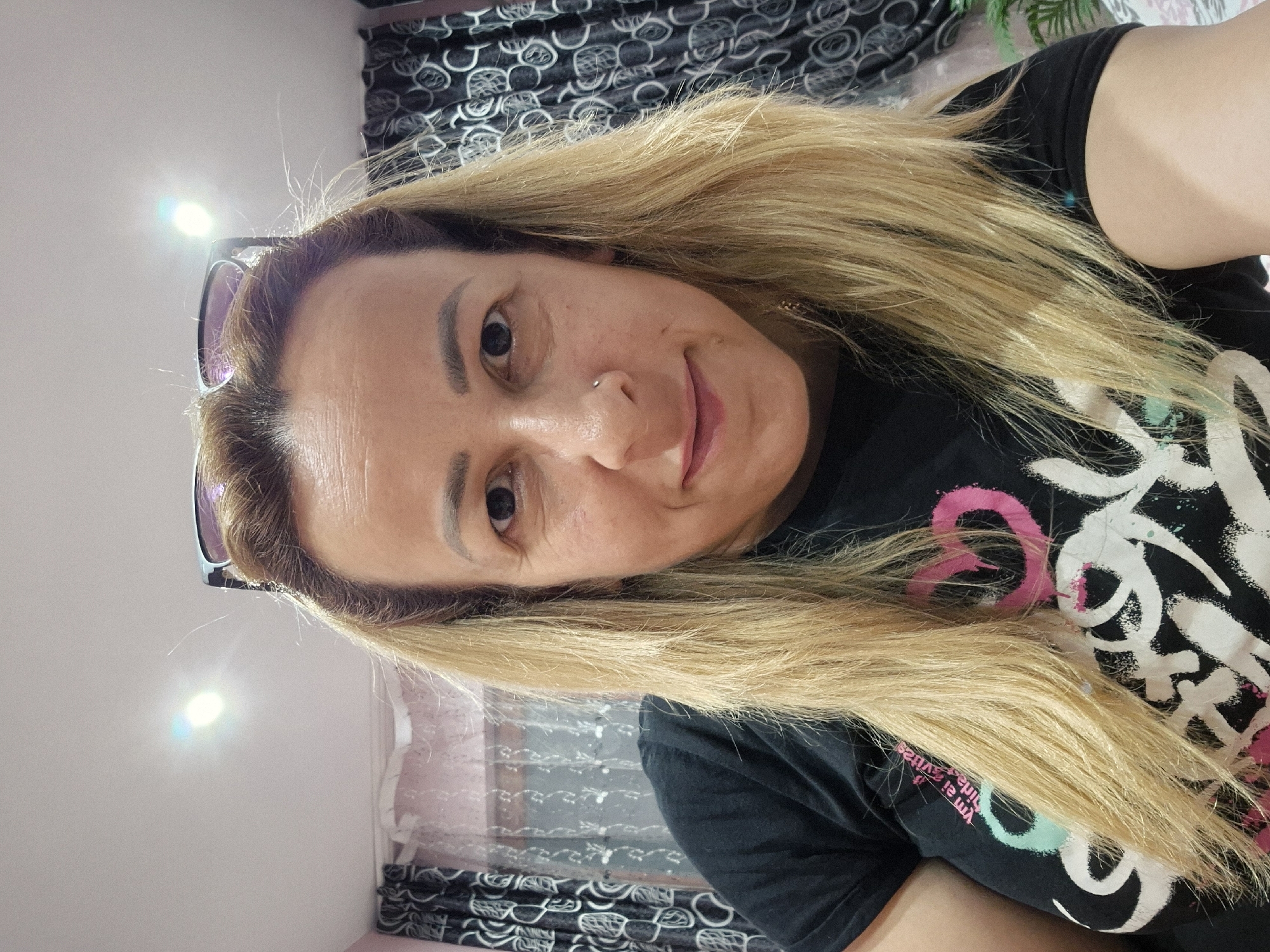 A close-up, front-facing selfie shows a woman with blonde hair, wearing glasses perched on top of her head and a black t-shirt with a colorful design. She has a nose ring and is looking at the camera with a slight smile. The background appears to be an indoor setting with curtains and ceiling lights visible. The lighting suggests it may be daytime, but the exact location is not specified.