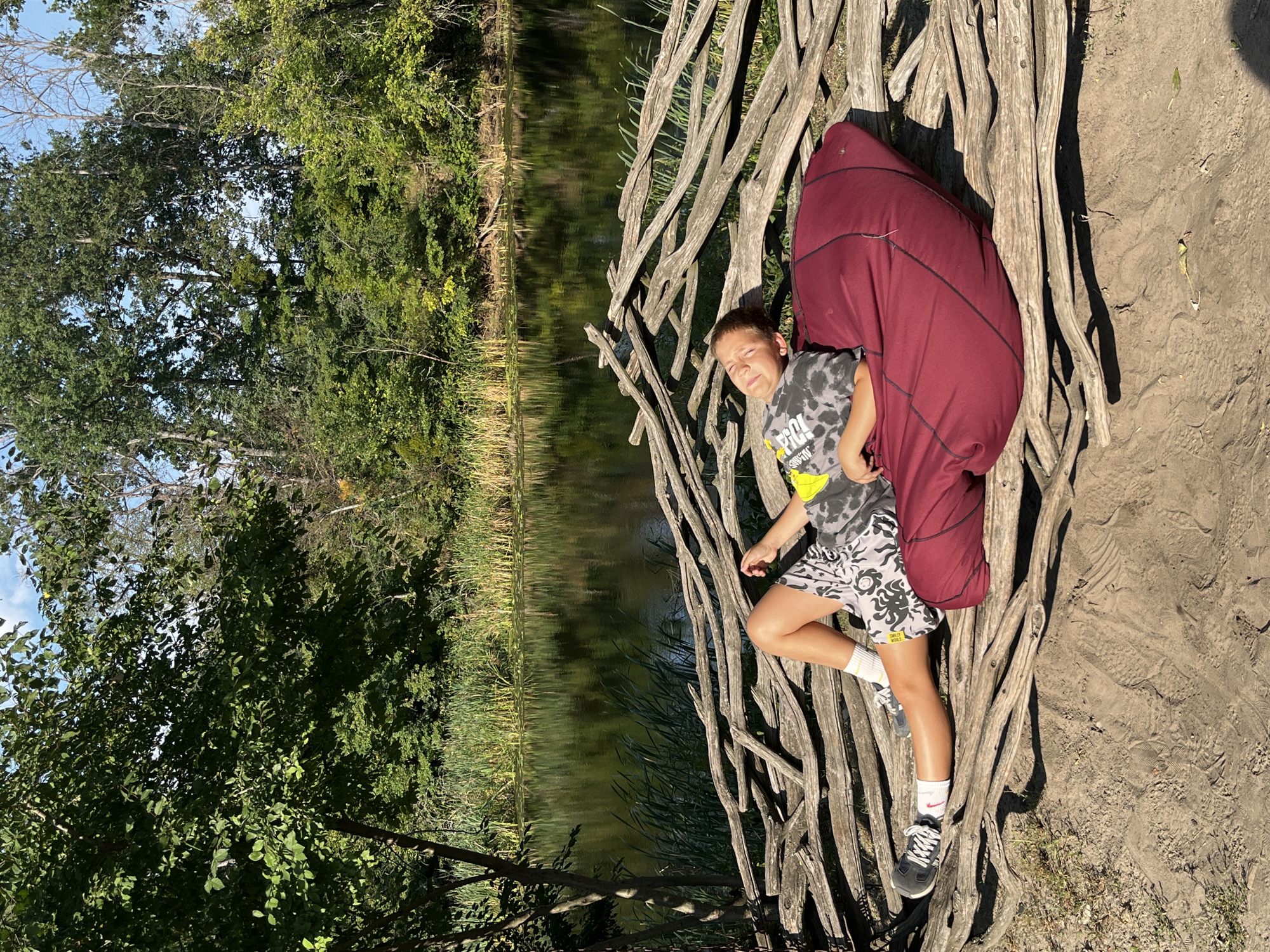 A young boy, wearing a tie-dye t-shirt and patterned shorts, is relaxing on a maroon beanbag chair placed on a rustic wooden structure made of interwoven branches. He is lying on his back with his eyes closed, enjoying the sunlight. The wooden structure is positioned in front of a body of water with tall reeds growing along its edge. Behind the water, a dense forest with lush green trees creates a backdrop. The ground in the foreground is sandy and appears to have footprints. The scene is set in the city of Korobiv Khutir, Ukraine, during the daytime.