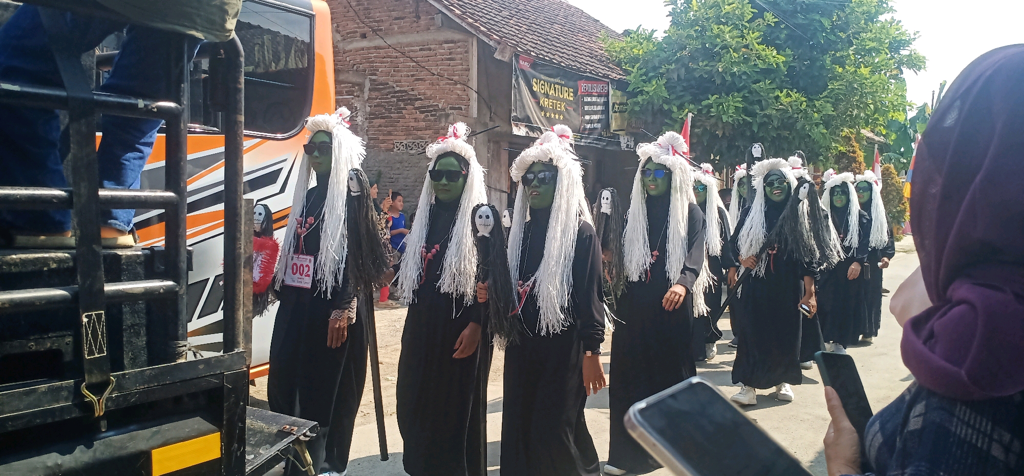 In Kincang, Indonesia, a group of individuals in costume participates in a parade. The participants, mostly female, are dressed in long black robes and wear distinctive white wigs and green face paint, resembling traditional Indonesian ghosts or spirits. Each person holds a prop, such as a doll-like figure with a white face and black hair, or a long pole. Some wear sunglasses, while others have red and white ribbons in their hair. A sign with "002" and text is visible on one participant's costume. Behind them, a bus with orange and white detailing is partially visible, as well as a brick building with a tiled roof. In the foreground, the edge of a truck is visible, suggesting the parade is moving along a street. The overall scene captures a vibrant cultural event within the Indonesian cityscape.
