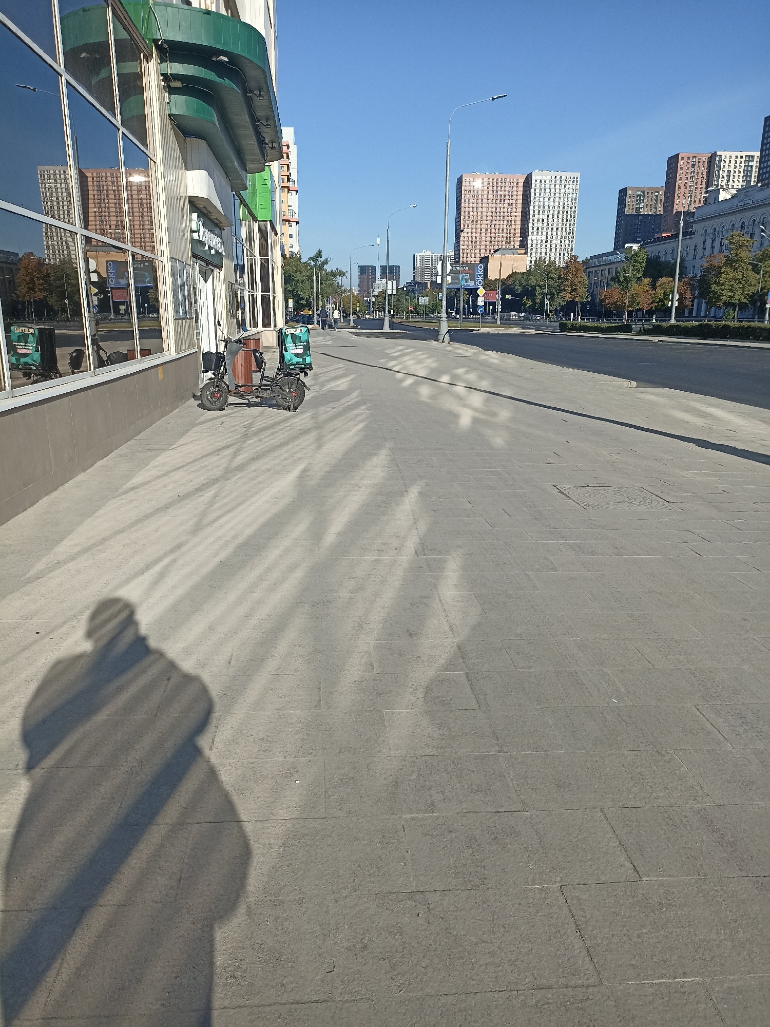 The image shows a wide, paved sidewalk in front of a modern building with large, reflective windows. Across the street, there is a road with traffic and several tall, colorful apartment buildings in the background. The sky is clear and blue, indicating a sunny day.
On the sidewalk, a shadow of a person holding a camera is visible in the lower left corner. To the right of the shadow, there is a parked electric scooter with two insulated delivery bags attached to it. The bags are teal and white, with some text and logos. The building on the left has green architectural elements and displays reflections of the buildings across the street. The sidewalk is made of light-colored rectangular paving stones, and there are long, striped shadows cast across it, likely from trees or overhead structures.
The scene is likely located in Moscow, Russia, as indicated by the architecture and the Cyrillic text visible on some of the reflections in the building windows, which appear to be advertisements or signage in Russian. The overall impression is of an urban environment on a clear, bright day.