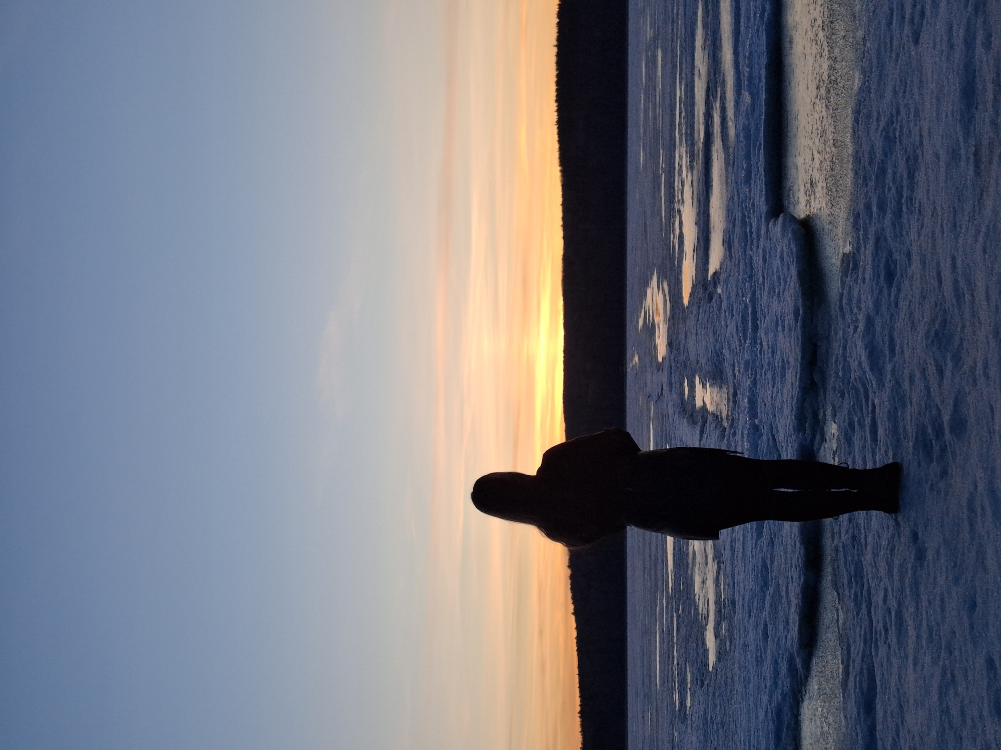 The image captures a solitary figure silhouetted against a dramatic sunset or sunrise over a frozen landscape in Chaikovsky, Russia. The composition, originally oriented vertically, depicts a wide horizontal expanse when viewed correctly.
In the foreground, a vast expanse of textured, frozen water, likely a lake or river covered in snow and ice, stretches across the right and lower portions of the frame. The ice appears uneven, with visible ripples and patches that reflect the colors of the sky.
A lone individual stands on this frozen surface, silhouetted in dark tones against the bright horizon. The person, positioned slightly to the right of the center, faces away from the viewer, seemingly observing the scene. Details of their clothing are indistinct due to the silhouette, but they appear to be wearing a coat and trousers.
The left and upper parts of the image are dominated by a clear sky, transitioning from a soft, pale blue at the top to vibrant oranges, yellows, and pinks closer to the horizon. This gradient suggests either a sunrise or sunset. A dark, narrow band of land, likely a treeline or distant shoreline, creates a sharp horizon line, separating the colorful sky from the frozen foreground.
The overall scene evokes a sense of tranquility and awe at the natural beauty of the cold landscape. The time of day is clearly either dawn or dusk, casting long shadows and creating the striking silhouette effect. No text is visible in the image.