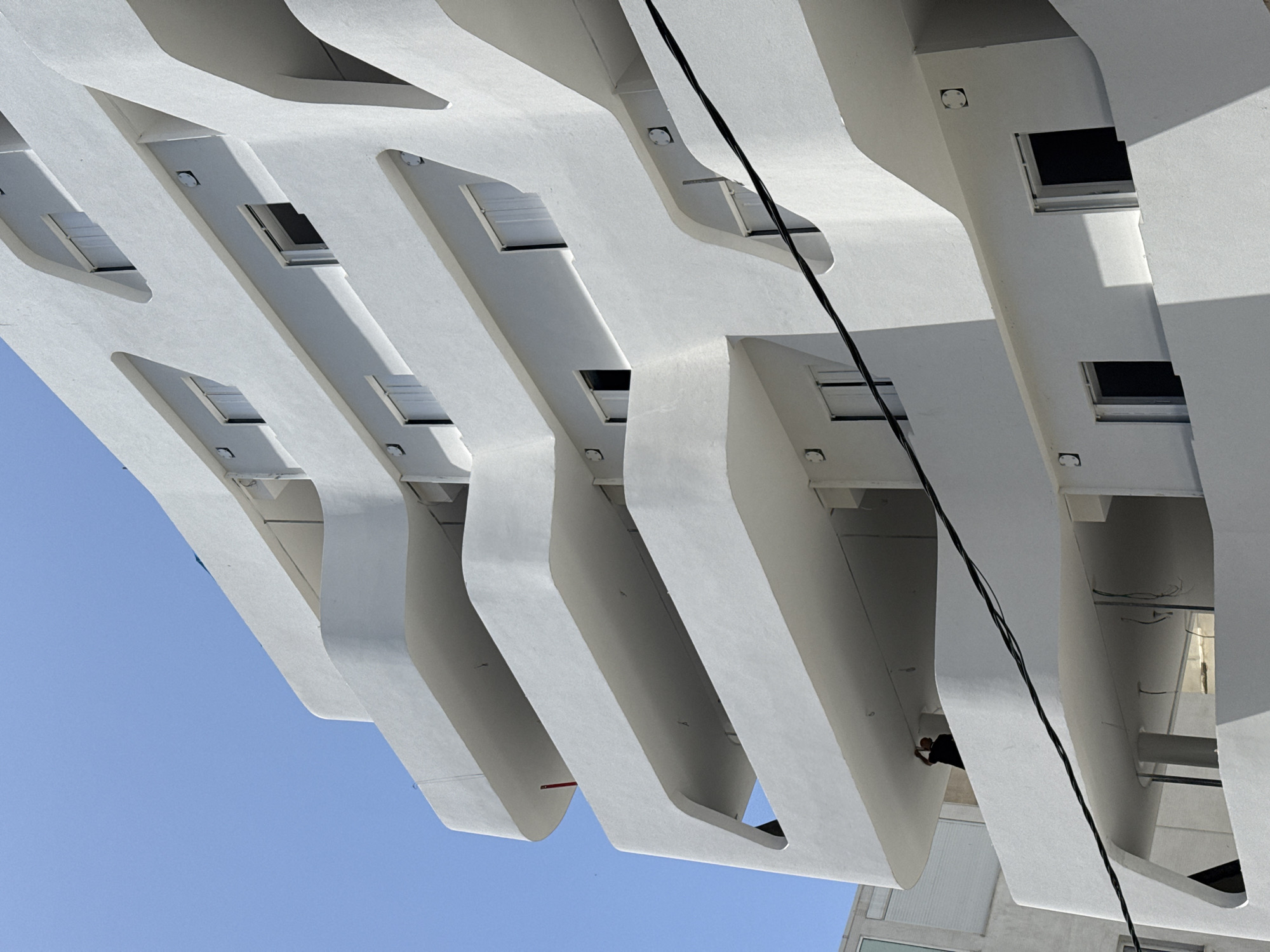 The image depicts a section of a modern building exterior in Tel Aviv, Israel. The building's design features repeating, elongated, white geometric protrusions with rounded edges, creating a rhythmic pattern against a clear, pale blue sky.
Small, rectangular window openings are integrated into the underside of each protrusion, adding depth and visual interest. A thick, black electrical cable runs diagonally across the frame, intersecting with the architectural elements.
In one of the openings, a person wearing dark clothing is visible, appearing to be working on or inspecting something within the structure. Another structure that is probably adjacent to the main one is visible in the bottom right corner of the frame.