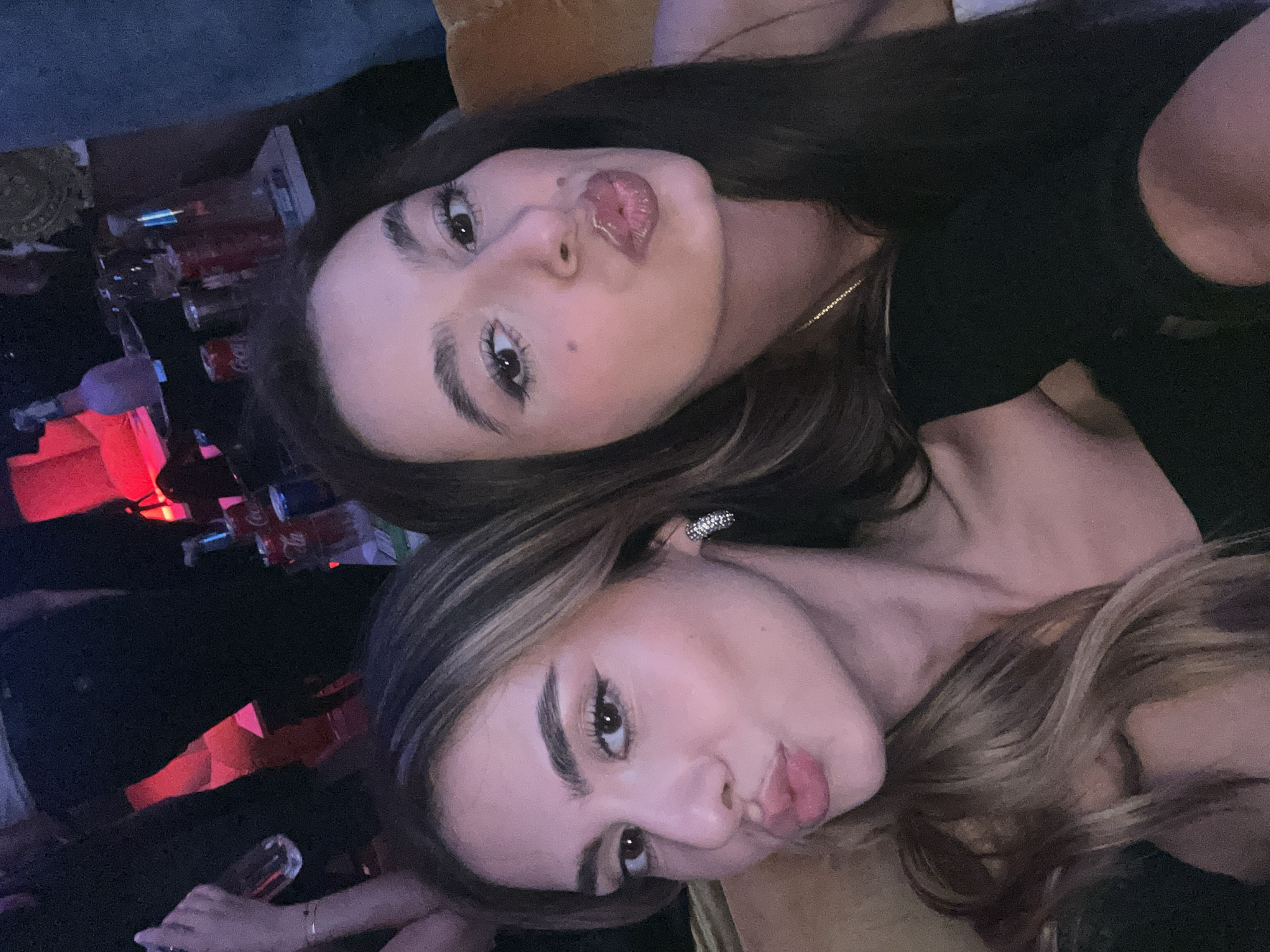 Two young women are posing close together, looking at the camera. The woman on top has long, dark hair and is wearing a black top. Her lips are pursed in a kissing expression. The woman below has long, wavy blonde hair and is also making a kissing expression. In the background, there is a table with drinks and a red light source. The image appears to be taken at night in a dimly lit bar or club. There is no indication of the location being in Pleven, Bulgaria.