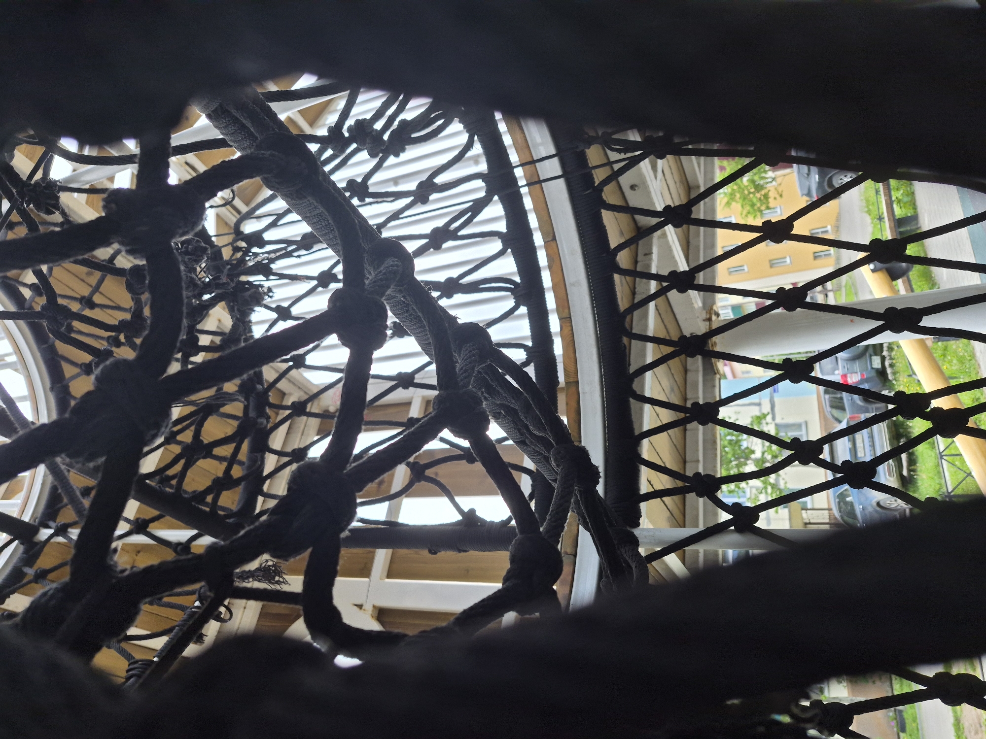 The image, taken in Uglegorsk, Russia, offers a low-angle, interior perspective of a rope structure, possibly a climbing net or hammock. Thick, dark ropes are intricately woven and knotted, forming a complex lattice. The ropes dominate the foreground and mid-ground, obscuring much of the background. Through the openings in the ropework, hints of an outdoor environment are visible, including buildings, parked cars, and green grass. The lighting suggests an overcast or shaded day. There are no people visible in the frame.