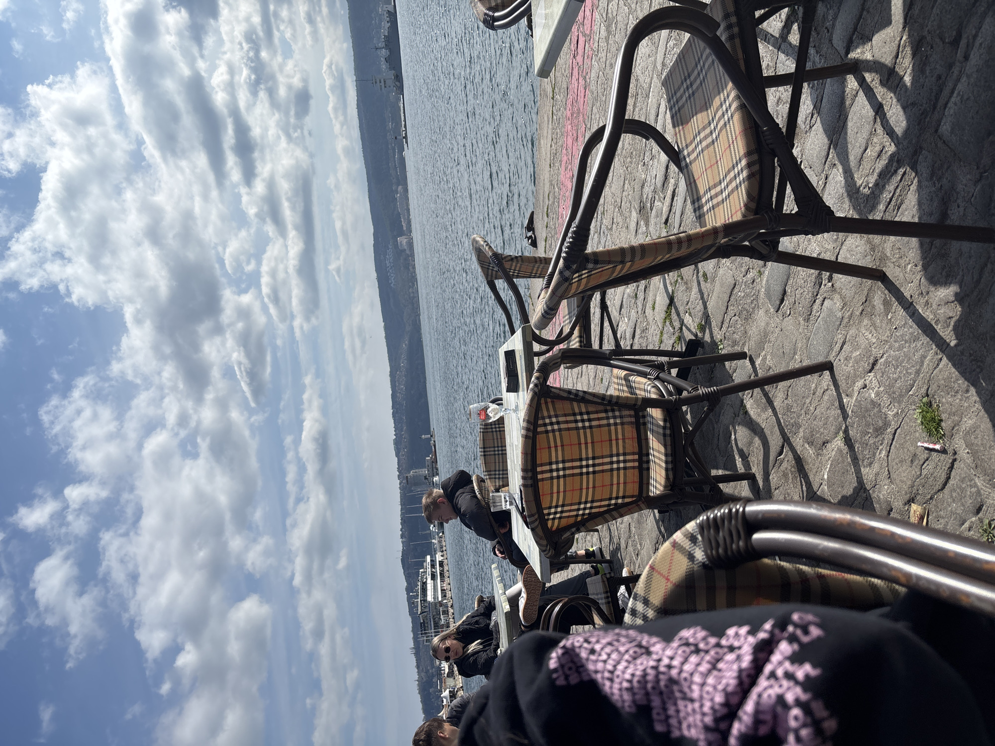 The image depicts an outdoor seating area in Varna, Bulgaria, situated near the coast. Several tables and chairs with a distinctive plaid pattern are arranged on a stone-paved surface. Three people are visible at the tables; one is seated with their face clearly visible, while the others are partially obscured. In the background, there is a body of water, likely the Black Sea, with land visible in the distance. The sky is partially cloudy.