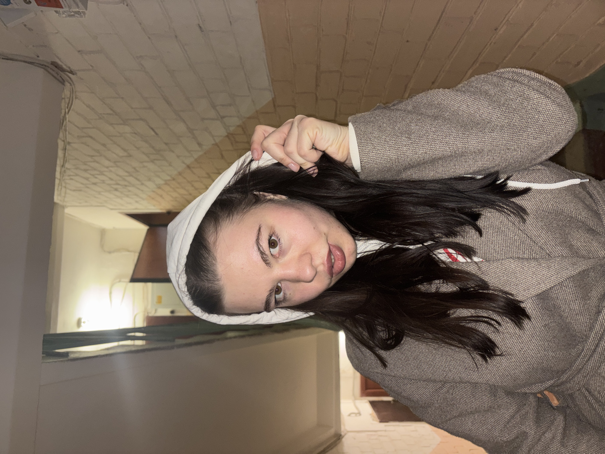 The image is an indoor portrait of a young woman wearing a hooded jacket. The hood is white, while the jacket appears to be a grey or brown tweed material. She is holding the hood with one hand. The background includes what appears to be a kitchen area with white cabinetry and what seems to be a brick pattern on a nearby wall. While it may resemble a location in the city of Perm, Russia, I cannot definitively confirm this based on the provided image alone.