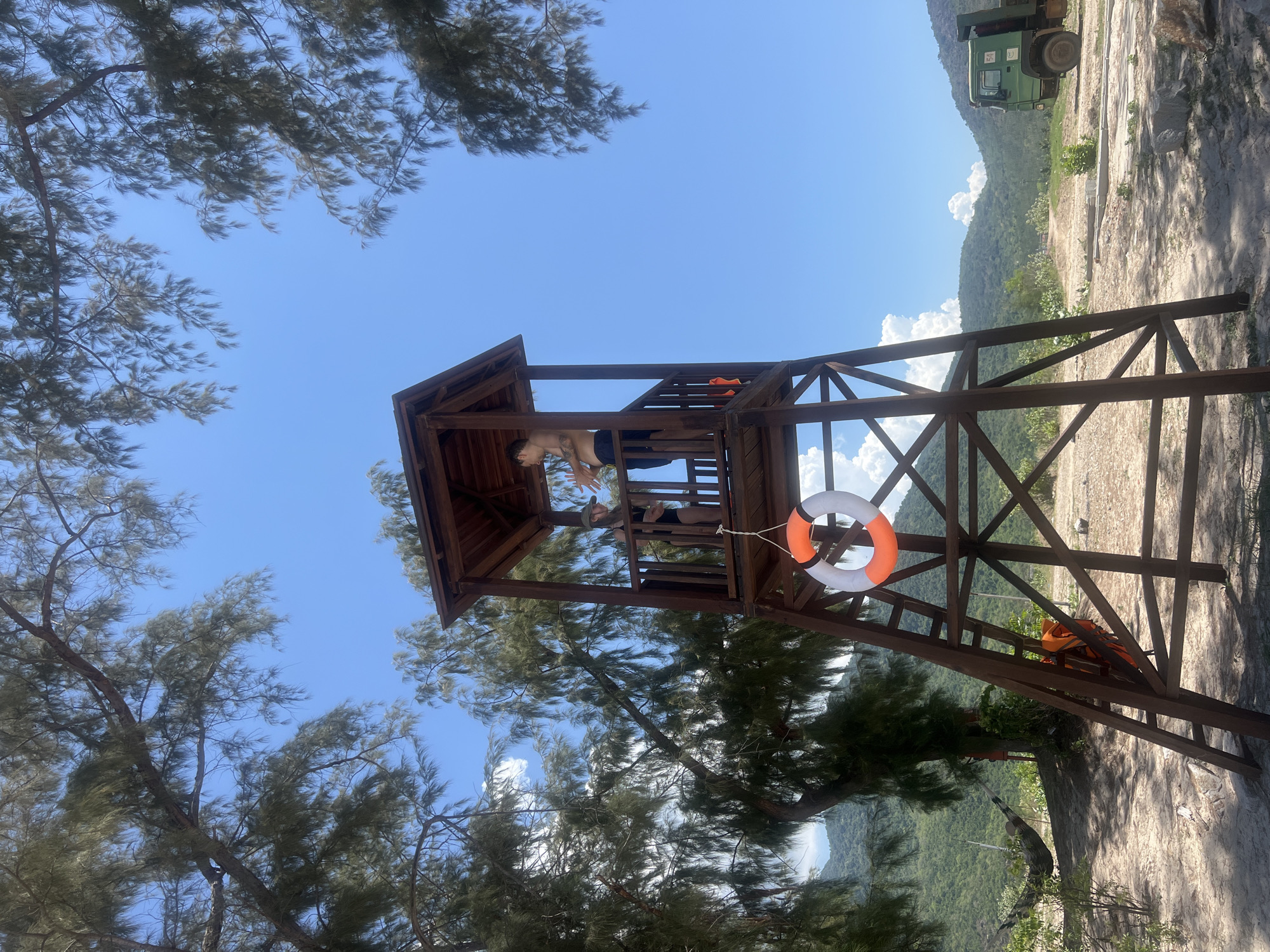 In Cong Hai, Vietnam, a wooden lifeguard tower stands on a sandy beach beneath a bright blue sky with scattered white clouds. Two individuals are visible on the platform of the tower. One, a person wearing a dark swimsuit and a green hat, is looking out. The other, a shirtless individual with tattoos on their arms, is facing the first person. An orange and white life preserver hangs from the tower's railing. To the left of the tower, a hammock is strung between two trees, and an orange life vest is attached to the tower's ladder. In the background, lush green hills rise, and a green truck with the number "54" visible on its side is parked on the sandy terrain. The scene captures a coastal environment with elements of beach safety and leisure.