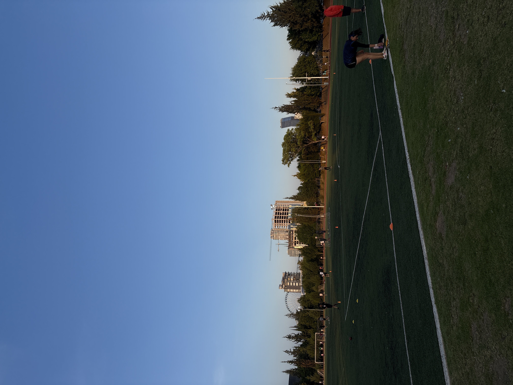 The image captures an outdoor sports field, likely for soccer or football, under a clear blue sky, set against an urban backdrop.
**Subjects:**
* **People:** In the foreground and mid-ground on the right side of the field, there are two prominent individuals. One person, wearing a dark blue long-sleeved shirt and dark shorts (with "UNDER ARMOUR" visible on the shorts), is bent over, seemingly stretching or adjusting footwear. They have dark, flowing hair and wear white sneakers with colorful accents. Further up the field, another person in a red shirt and dark shorts is standing. Several other smaller figures are distributed across the field in the distance.
* **Objects:** The field itself is covered in green grass, clearly marked with white lines indicating playing boundaries. Several orange and yellow training cones are scattered on the grass. Along the edge of the field, tall white poles, possibly for lighting or flags, are visible. In the background, beyond a treeline, numerous modern buildings are present, including one under construction with visible cranes. A large Ferris wheel stands out prominently among the buildings in the mid-distance.
* **Scene/Setting:** The scene is an athletic complex or public sports ground situated within an urban or suburban environment, bordered by trees and city structures.
**Activity or event taking place:** The individuals on the field, dressed in athletic wear and positioned near training equipment like cones, appear to be engaged in an athletic practice, training session, or warm-up.
**Notable details:**
* **Time of day:** The lighting suggests late afternoon or early evening, characterized by a clear, bright blue sky and soft, warm sunlight.
* **Weather:** The sky is completely clear and cloudless, indicating pleasant weather.
* **Location cues:** The distinctive urban skyline, including the Ferris wheel and specific buildings, suggests a particular location. Given the provided context, this scene is in Puebla, Mexico.
* **Visible text:** The brand "UNDER ARMOUR" is clearly visible on the shorts of the person in the dark blue shirt.