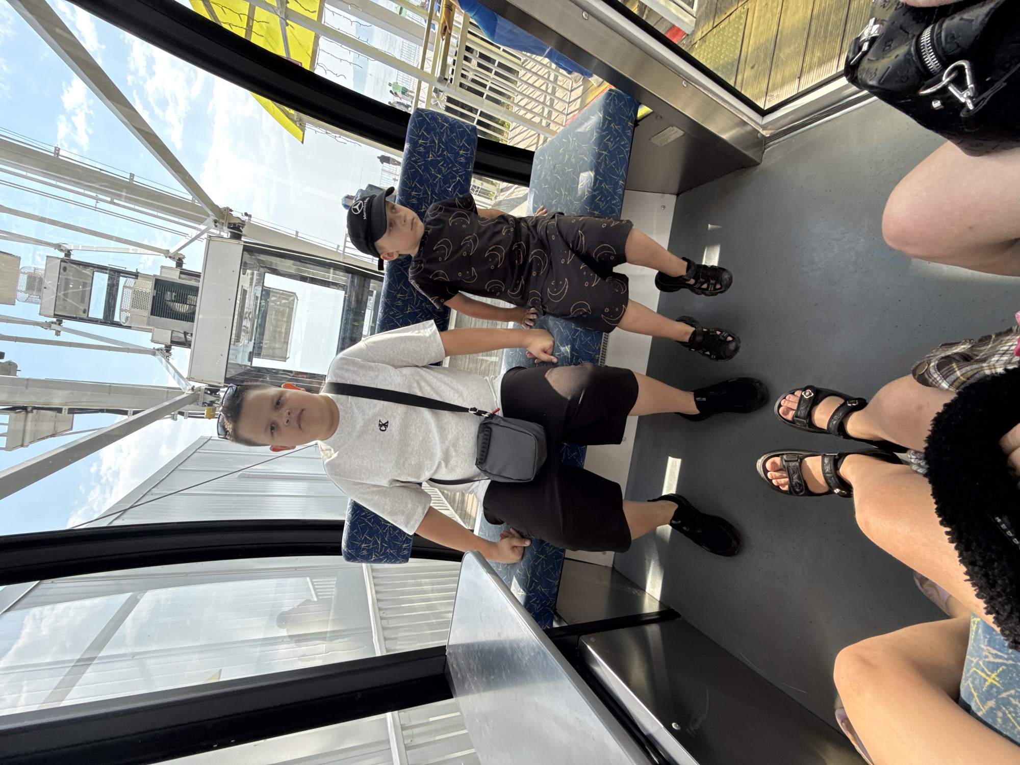 Two boys are seated inside a cabin of a Ferris wheel. The boy on the left, appearing older, wears a light gray t-shirt, black shorts, and black sneakers, with a small dark gray shoulder bag. The younger boy on the right wears a black t-shirt and shorts with a crescent moon pattern, a black baseball cap, and black sandals. They are seated on blue and white patterned benches. A metallic table is visible to the left of the older boy. The Ferris wheel structure and a body of water are visible through the cabin windows. The scene is set within the town of Listvyanka, Russia, a popular tourist destination on the southern shore of Lake Baikal.