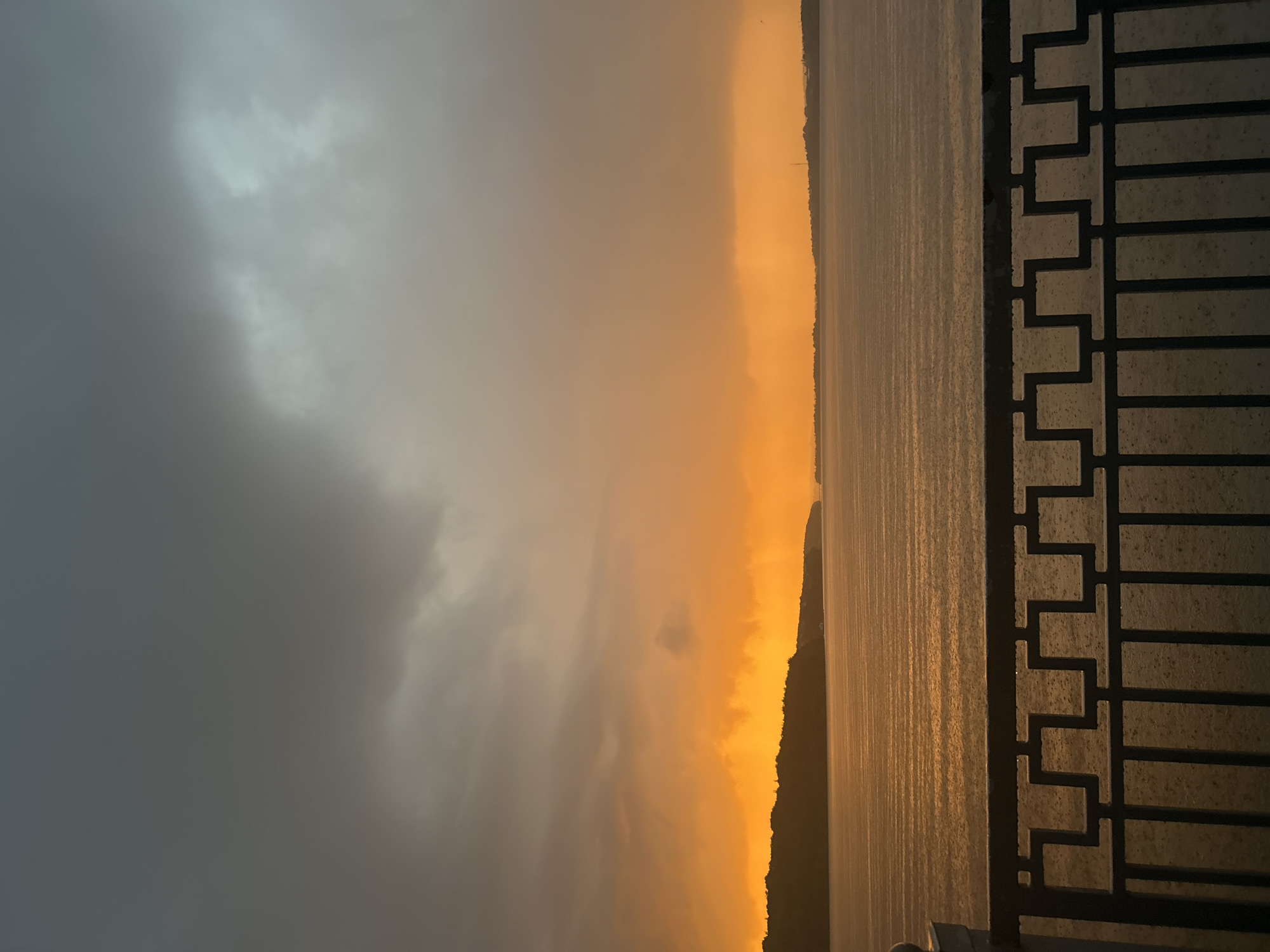 This is a photograph of a dramatic sunset over a body of water, likely Ternopil Lake, in Ternopil, Ukraine. The sky is a mix of dark, ominous clouds in the upper portion, transitioning to a vibrant, fiery orange and yellow glow near the horizon. The light from the sunset casts a warm, golden reflection on the rippling water, which appears to be experiencing rain. In the foreground, a dark metal railing with an ornate geometric pattern separates the viewer from the scene. The distant shoreline is silhouetted against the bright sky, with a faint line of trees and structures visible. The overall atmosphere is one of moody beauty, with the rain adding a dynamic element to the already striking sunset. There are no people or animals visible in the image.