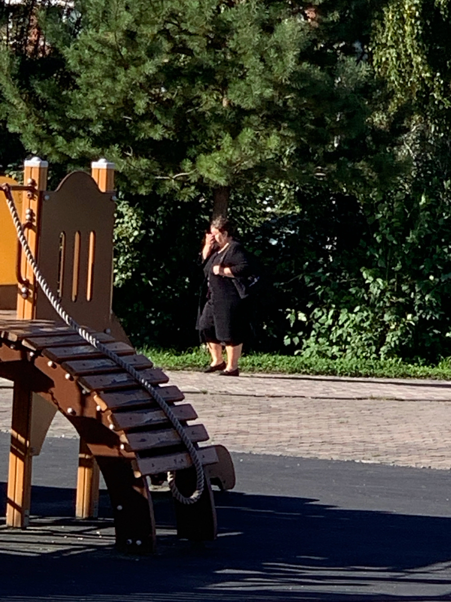 A medium-shot, eye-level photograph captures a woman walking on a paved path next to a children's playground. The woman, wearing a dark dress and black shoes, holds her hand to her face as she walks to the left of the frame. A rope ladder from the playground extends into the foreground. In the background, lush green trees and foliage dominate the scene, suggesting an outdoor park or recreational area in Prokopyevsk, Russia, on a sunny day.