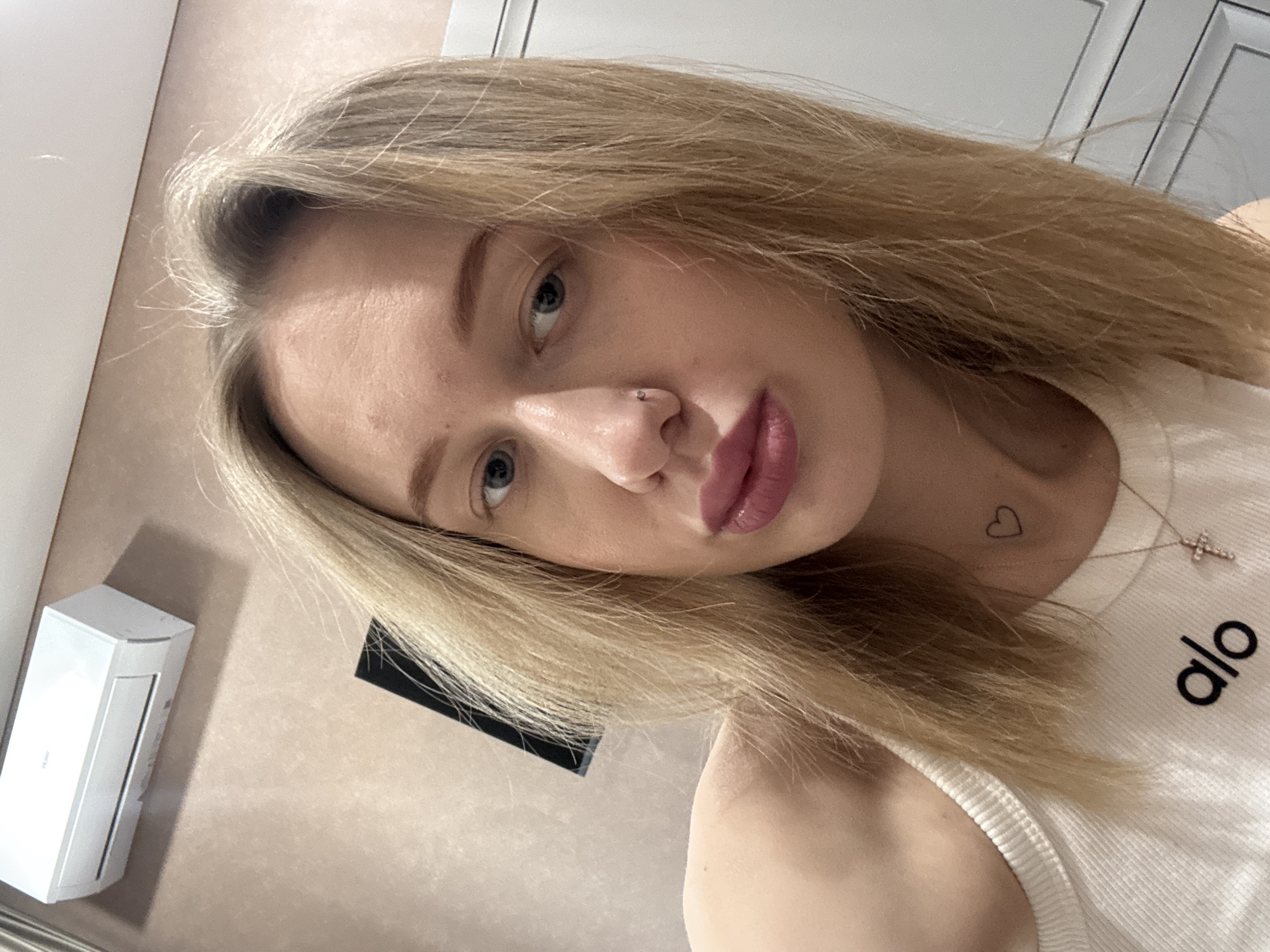 A blond-haired woman poses indoors. She is wearing a white tank top with "alo" printed on it, a necklace with a cross, and has a nose piercing and a heart tattoo behind her left ear. A white air conditioner unit is mounted on a beige wall. A black object is also affixed to the wall. A white door with panels is in the background. There is no information to suggest that this image was taken in Ovoshchnoy, Russia.