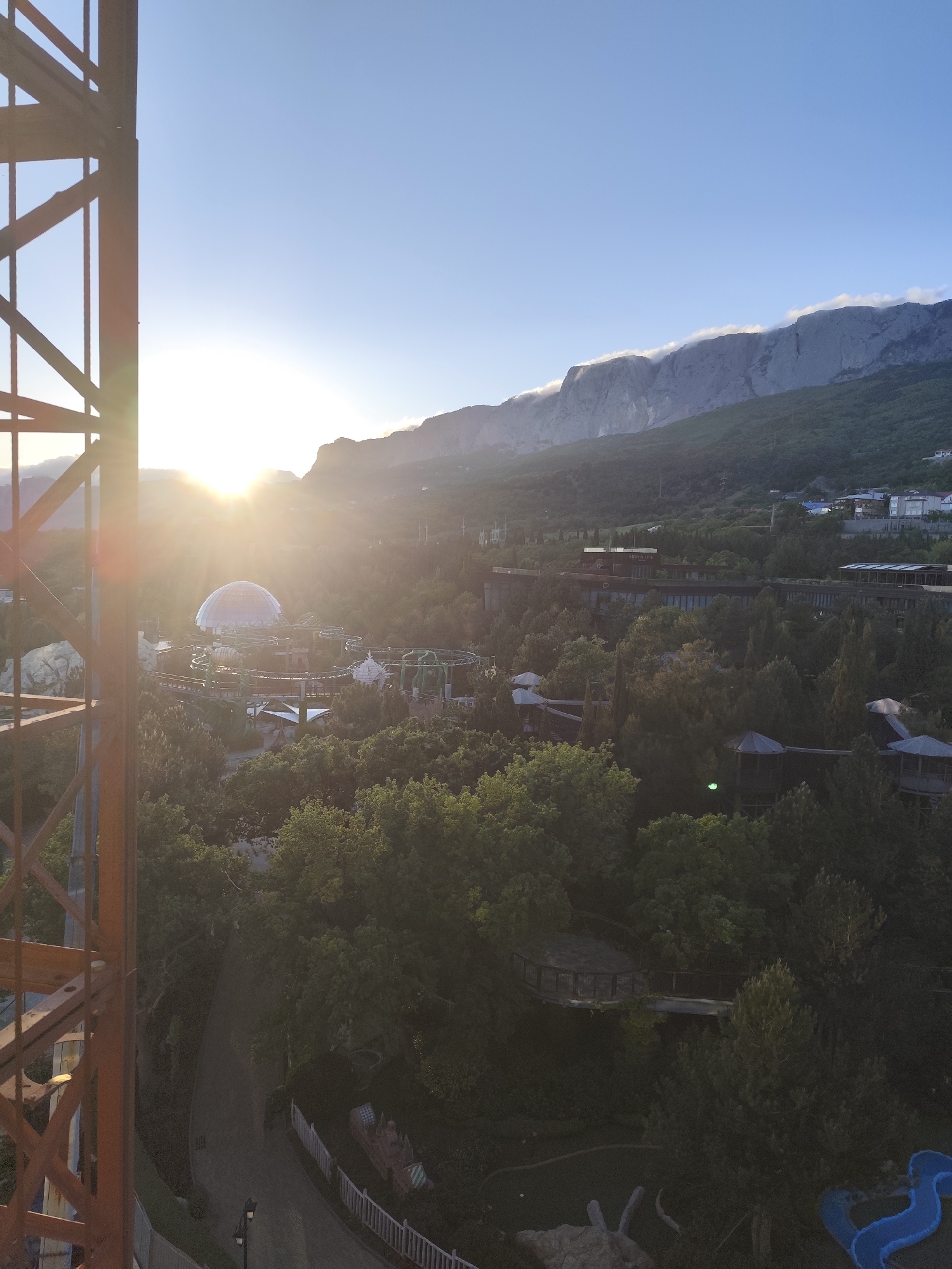 The image shows a landscape view in Симеизский поселковый совет, Ukraine, captured from an elevated perspective. In the distance, a mountain range stretches across the horizon, partially covered in clouds. Closer to the viewer, there is a theme park or amusement park with rides and a large dome-shaped building. The landscape is heavily vegetated with trees. A metal structure, possibly part of the construction of the elevated platform, is visible on the left side of the image. The sun is positioned in the upper left corner, creating a lens flare. Buildings are seen scattered within the trees and on the side of the mountains.