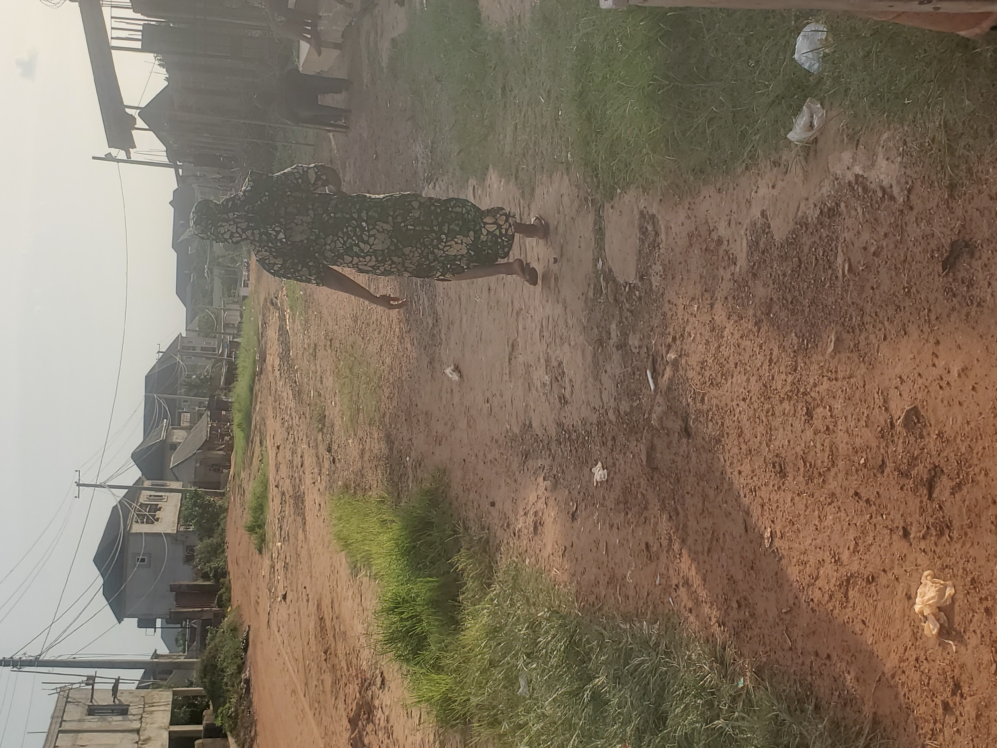 The image depicts a dirt road in Ukwu-Abwa, Nigeria. A woman wearing a patterned dress and head covering is walking along the road. The road is bordered by grass and vegetation, with houses and buildings visible in the background. Power lines and poles are also visible, suggesting the area is somewhat developed. Scattered pieces of trash are seen on the ground.