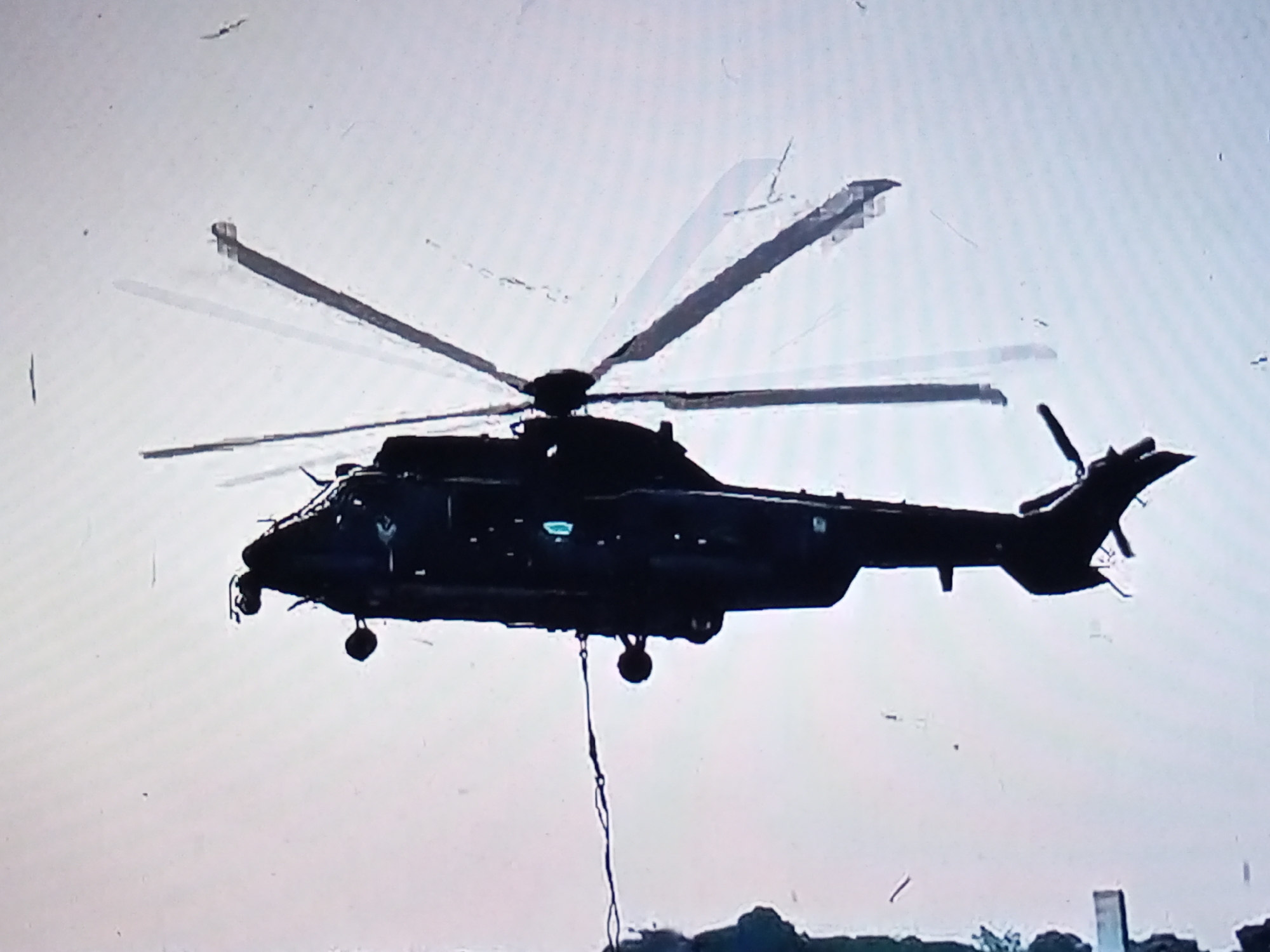 An aerial, side-view, black-and-white image captures a dark helicopter in flight against a hazy, light sky, with a faint, dark horizontal line at the bottom of the frame. The helicopter is silhouetted, with its rotor blades blurred due to motion, indicating rapid rotation. A dark cable hangs from the underside of the helicopter, trailing downwards. The overall image quality is low, with visible artifacts and a grainy texture, suggesting it may be a low-resolution capture or a photograph of a screen. The setting does not provide any specific visual cues to confirm the location as Banyudono, Indonesia.