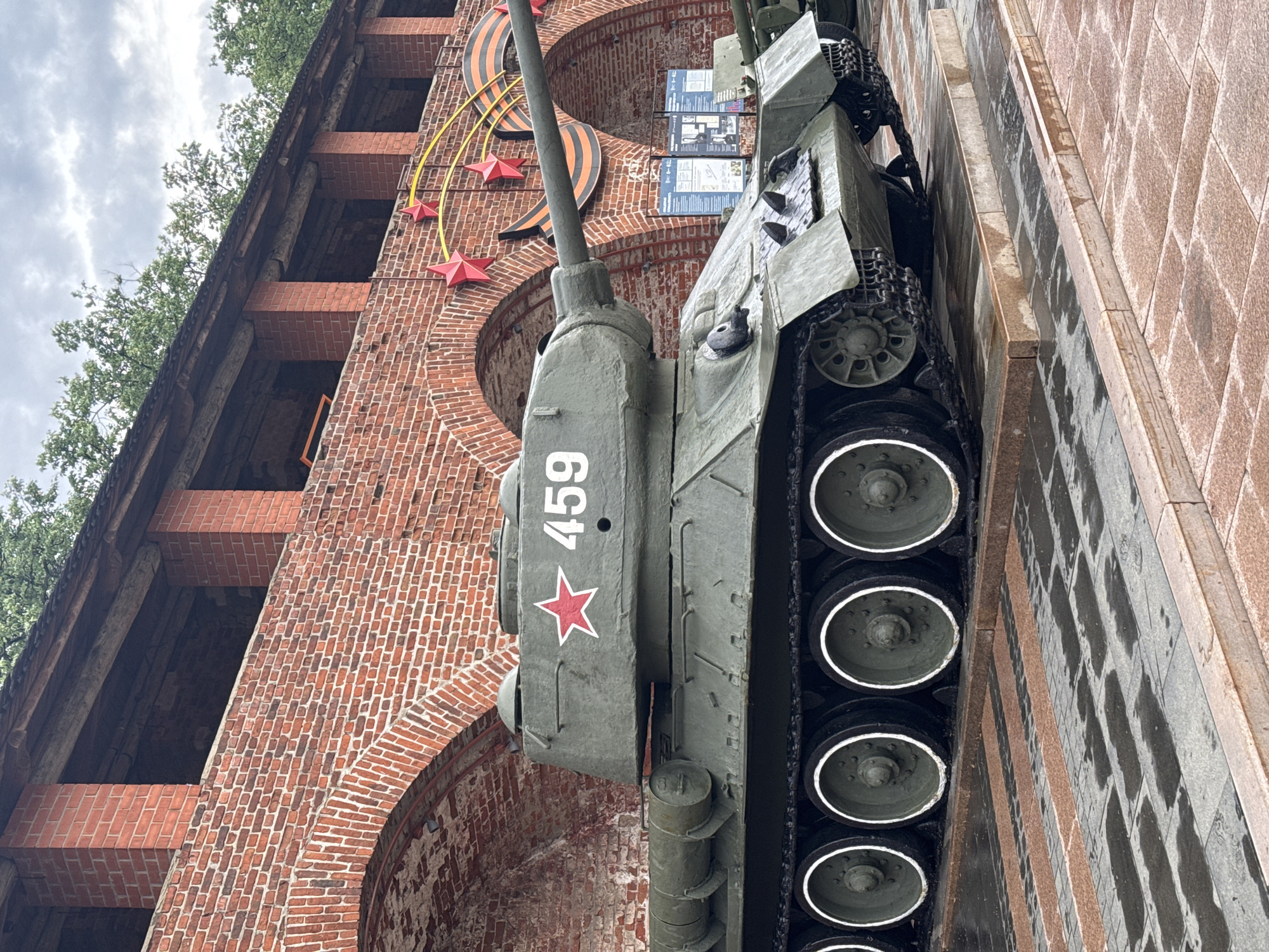 A Soviet-era tank with the number "459" and a red star painted on its turret is positioned on a stone platform beside a red brick building with arched openings. The tank's treads and wheels are visible, along with the gun barrel aimed upwards. The building is adorned with a large orange and black ribbon and red star decoration. The scene appears to be in Nizhny Novgorod, Russia.