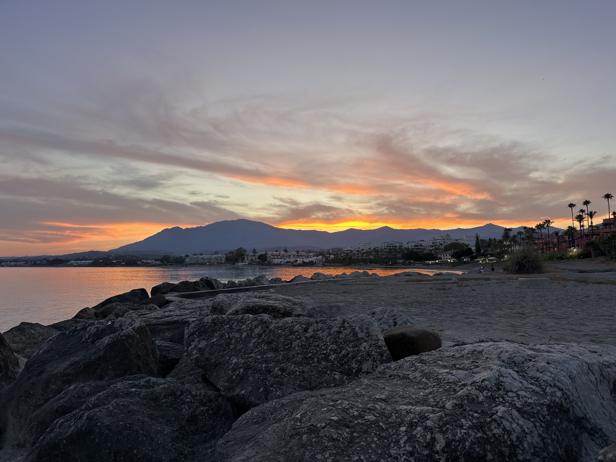 A serene coastal sunset scene unfolds in El Velerín, Spain. The sky is a canvas of soft purples, pinks, and vibrant oranges, with wispy clouds stretching across the horizon. In the distance, a range of mountains forms a dark silhouette against the glowing sky, with a town nestled along the coastline. The water is calm and reflects the warm hues of the sunset. In the foreground, large, dark rocks line the shore, interspersed with a sandy beach. A few palm trees are visible along the coastline, adding to the tropical ambiance. The overall impression is one of peaceful tranquility.