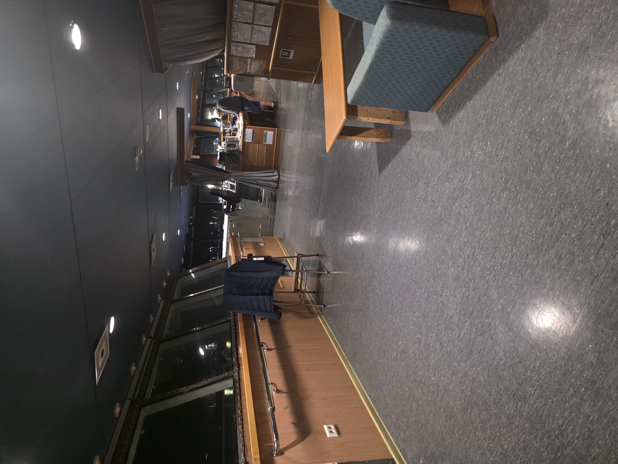 The image depicts the interior of a ship's bridge, likely at night, given the darkness visible through the large windows. The bridge is functional and appears to be in use. The floor is a dark gray linoleum. A dark blue jacket is draped over a metal stool positioned against a wooden paneled wall. A metal handrail runs along the wall. To the right, a wooden bench with blue upholstered seating is visible. In the background, a person in casual clothing is standing at a console or desk, appearing to be working. The ceiling is dark blue with several recessed lights. The overall impression is one of a working environment on a vessel. The image does not provide any specific visual cues to pinpoint its location to Brindisi, Italy.