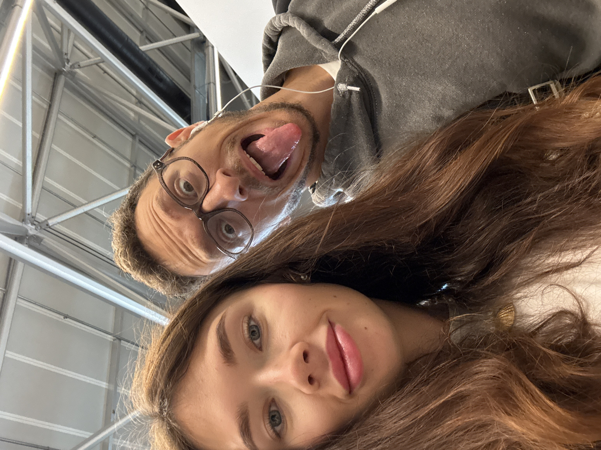 A selfie taken from a low angle, looking up at two people and the ceiling above. A woman with long brown hair is in the foreground, her face tilted slightly. She has fair skin, blue eyes, and pink lipstick. Behind her is a man wearing glasses, with his tongue sticking out and mouth open in a silly expression. He is wearing a gray hooded sweatshirt and has earbuds in his ears. The background is a ceiling with a metal grid structure and pipes, suggesting an indoor setting, possibly an airport or a similar public space. The location appears to be an airport.