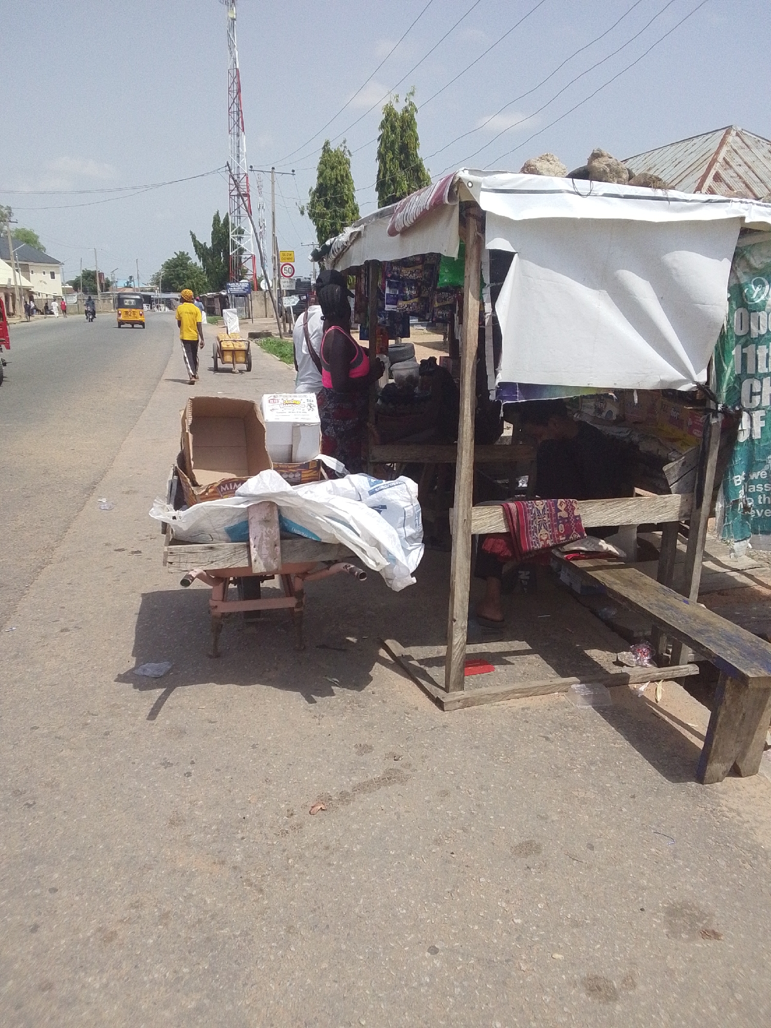 This image captures a street scene in Jalingo, Nigeria. A roadside stall, likely selling various goods, is set up with a white tarpaulin roof supported by wooden poles. In the foreground, a weathered wooden cart or trolley holds cardboard boxes and a large white plastic bag.
Several people are visible around the stall. A person in a bright yellow shirt and dark pants walks away from the camera, pulling a similar cart. Behind them, a yellow tricycle, possibly a form of local transport, is on the road. Closer to the stall, a person in a pink top and patterned skirt stands, appearing to interact with the stall owner or a customer. Another individual is seated within the stall, partially obscured.
The scene suggests a busy market or commercial area. The weather appears to be bright and sunny, casting distinct shadows on the asphalt road. A tall telecommunications tower stands prominently in the background, along with power lines crisscrossing the sky. A road sign indicating "SLOW DOWN 50" is visible on the left side of the road. The overall atmosphere seems typical of an active day in a Nigerian town.