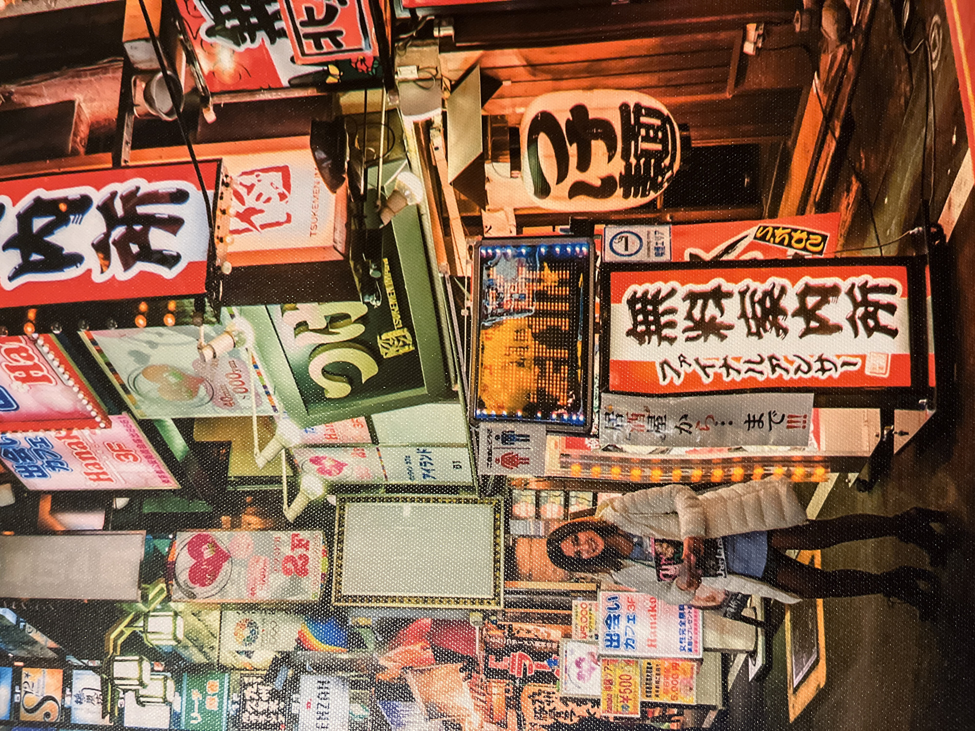 The image depicts a street scene at night, heavily adorned with illuminated Japanese signs. In the foreground, a woman stands with her back to the camera, facing a wall of signage. She is wearing a white puffer coat, a blue top, black tights, and black heeled boots. She is holding a book or magazine in her hands. The background is a dense arrangement of neon and kanji-emblazoned signs, typical of a bustling entertainment district. Some signs are in red and white, others in various colors, and some are lit with warm yellow lights. A large, white paper lantern with black Japanese characters hangs prominently on the right side of the image. The ground appears to be wet, reflecting the bright lights of the signage. The overall atmosphere is one of vibrant urban nightlife.