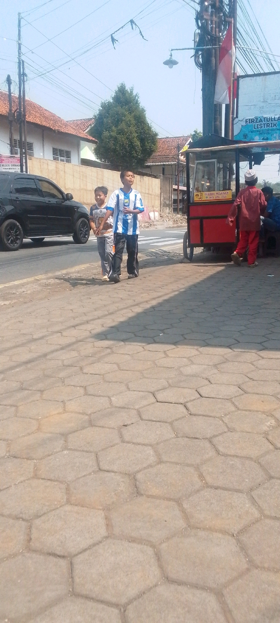 Two young boys, one in a blue and white striped soccer jersey and black pants, the other in a patterned shirt and gray pants, walk on a hexagonal paver sidewalk in Sirnagalih, Indonesia. A black SUV is parked on the street to the left. A red food cart with a white awning and a sign reading "TERIMA PESANAN 0878 1857 1735" is situated on the right side of the sidewalk. A man in a red shirt and white cap stands near the food cart, facing away from the camera. Another person, partially obscured, sits on a bench to the right of the food cart. Overhead wires and a street lamp are visible against a clear blue sky. Buildings with red tile roofs line the background.