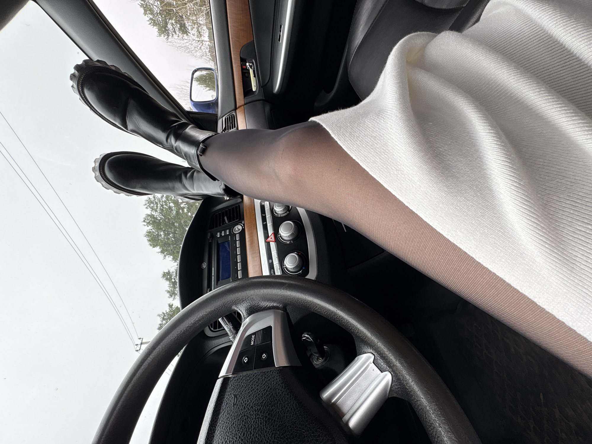 The image depicts the interior of a car, with a steering wheel in the foreground. A woman's legs, clad in sheer black stockings and black boots, are resting on the dashboard or front window. The woman is wearing a white sweater. The car's dashboard, center console, and rearview mirror are visible. Through the front window, one can see a wintry scene of trees, likely near Chaikovsky, Russia based on contextual information.