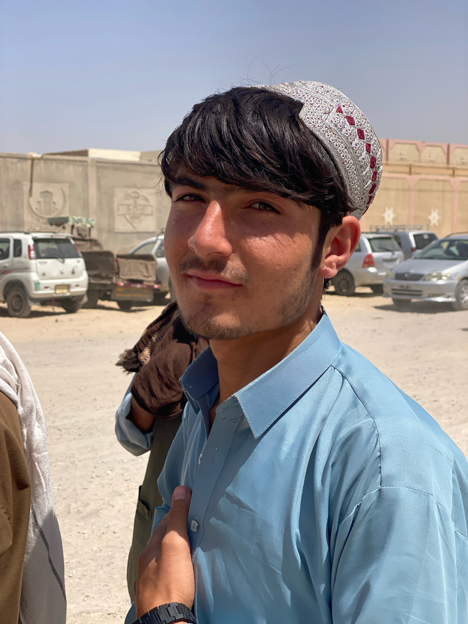 In the city of Kandahar, Afghanistan, a young man is captured in a medium close-up shot outdoors, under a clear, bright sky. He is looking directly at the camera, with a slight, almost imperceptible smile. He is wearing a light blue collared shirt and a distinctive white and red patterned cap. His hair is dark and neatly styled. His right hand is resting on his chest, near his collarbone.
The background suggests an urban or semi-urban environment, with sandy ground and a low concrete wall or barrier. Several vehicles are visible in the distance, including cars and what appear to be small trucks or utility vehicles, parked in a lot or along a road. The overall lighting indicates it is daytime, with strong sunlight casting shadows. The image has a slightly informal, candid feel.
