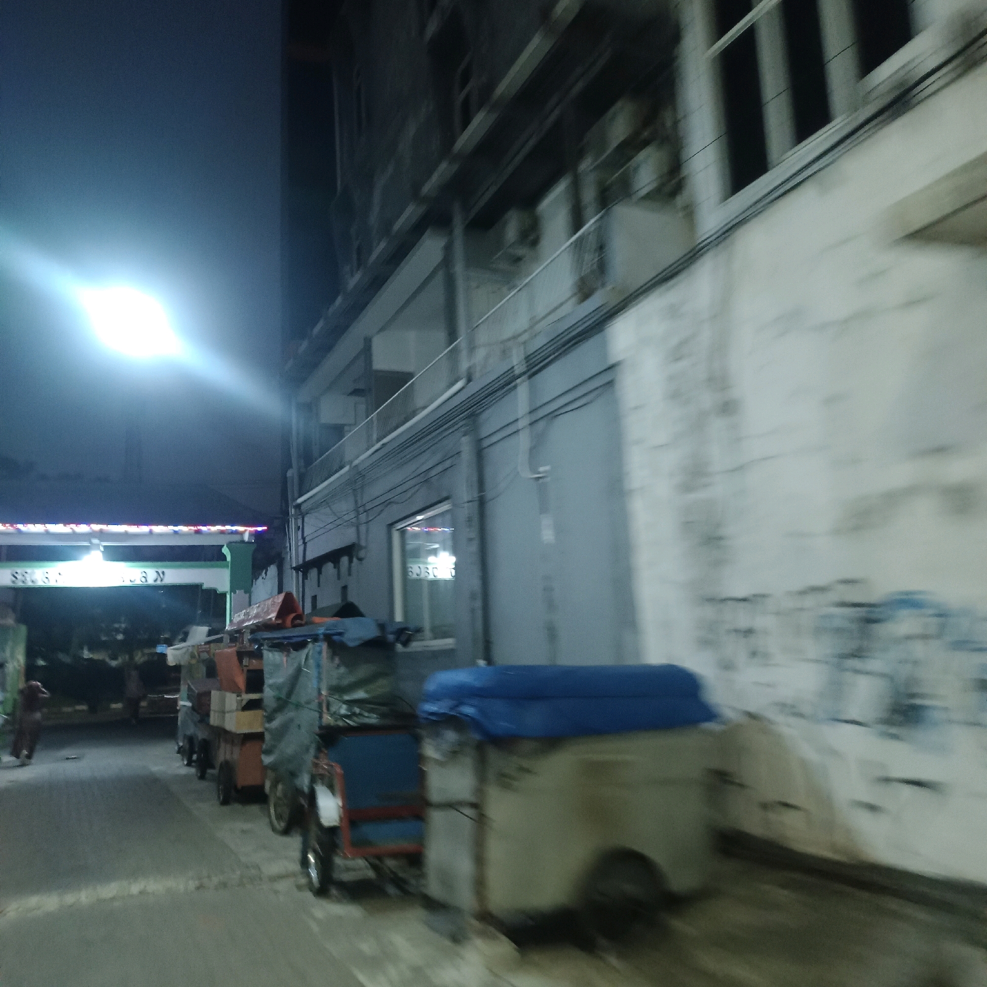 The image shows a street scene at night in Surabaya, Indonesia. A bright light is visible in the upper left corner, potentially a street lamp or a vehicle's headlights. In the center of the image, there are several carts, some of which appear to be food vendors, with items stacked on them. One cart is covered with a blue tarp. To the right of the carts, a building with a light-colored, textured wall is visible. The building has several windows and is partially obscured by wires. In the background, there is another building with darker windows and architectural details. The overall impression is of a street or market area at night, with a slightly blurred quality suggesting movement. There are a few indistinct figures of people present, but their actions are not clear. The street itself appears to be paved.