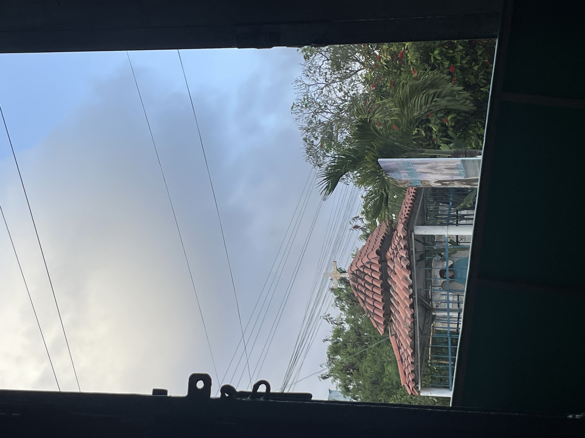 The image, taken from an indoor vantage point in Porlamar, Venezuela, shows an outdoor view through a window or doorway. Above is a cloudy sky. A building with a tiered, terracotta-tiled roof is visible, surrounded by lush green trees and other foliage. Utility wires cut across the sky. A small white cross is fixed atop the building. A partial view of what could be a poster or advertisement is present, attached to the gazebo. The framing elements of the window or doorway are visible, including dark hardware at the bottom.