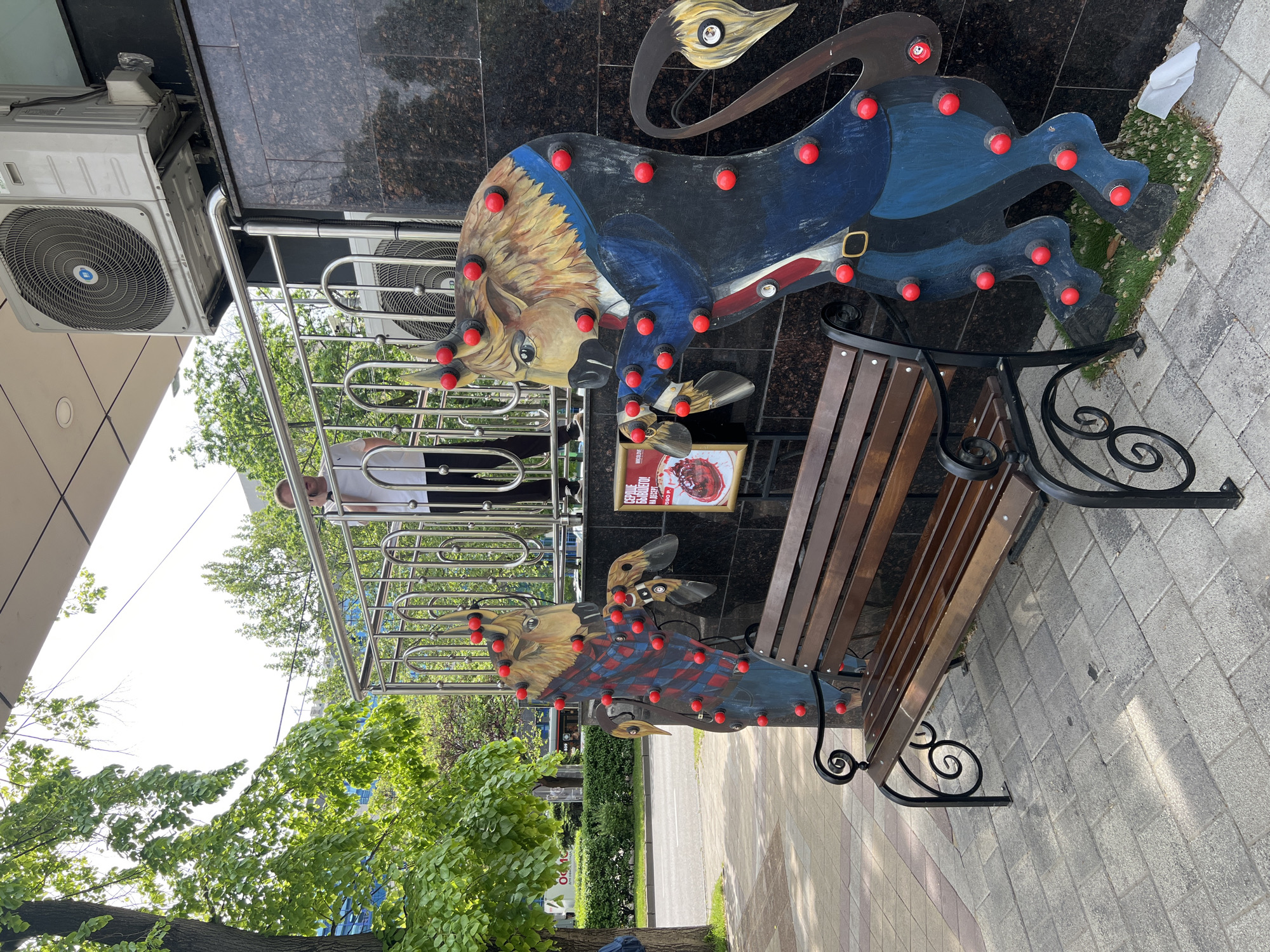 The image shows a public space in Krasnodar, Russia. A park bench with ornate metal supports is the central object. The bench is flanked by two large, painted wooden figures resembling lions or bulls, adorned with red light bulbs. The figures appear to be integrated into the design of the bench. Behind the bench is a building with a dark tiled exterior, with a metal railing and a person visible on an upper level. An air conditioning unit is mounted on the building's exterior. A poster is attached to the wall between the wooden figures. A tree is visible to the left of the bench, providing some greenery. The ground is paved with light-colored bricks.