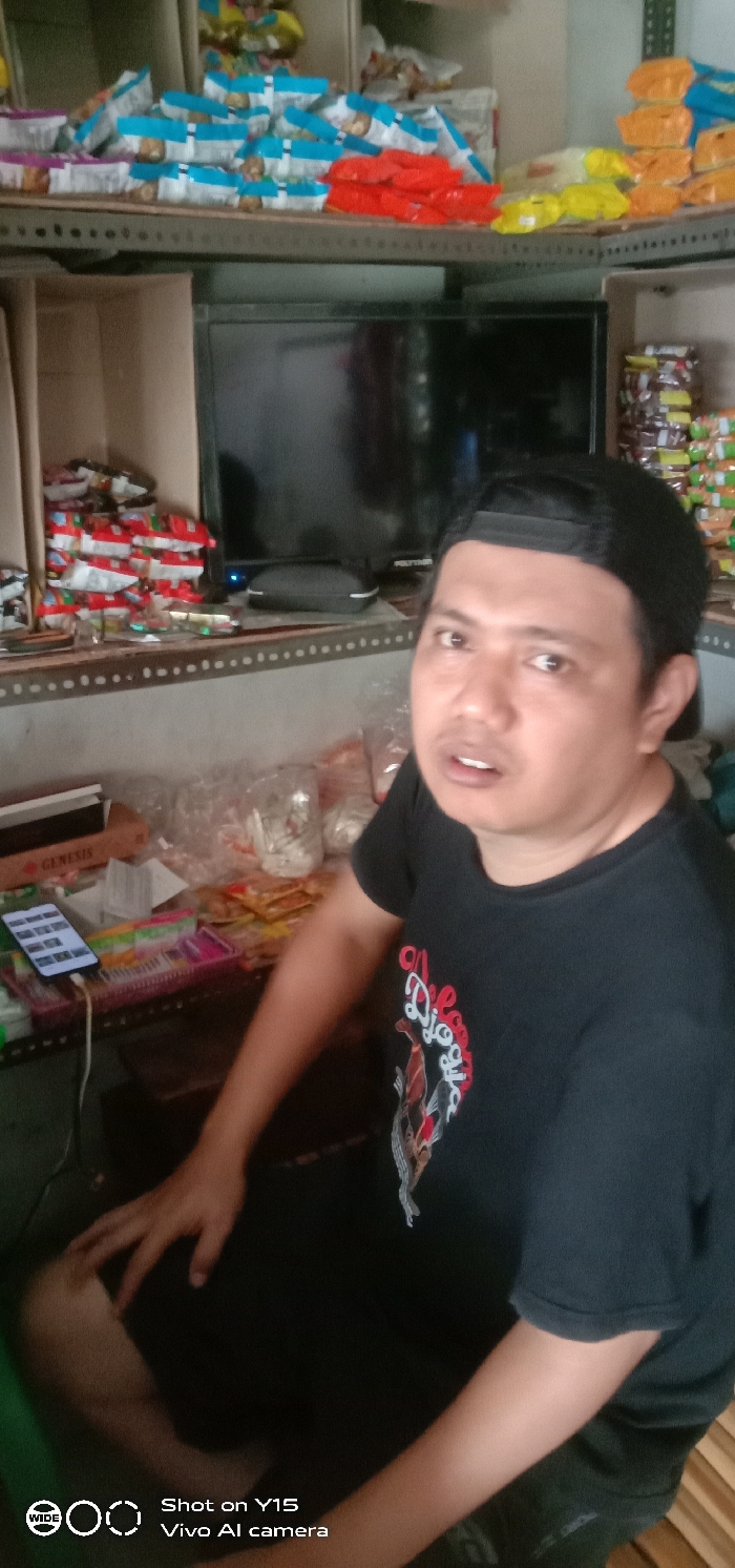 The image depicts the interior of a small retail establishment or "warung" located in Sebaung, Indonesia. A single adult male, appearing to be of Southeast Asian descent, is seated and positioned prominently in the lower right and center of the frame, looking directly at the camera with his mouth slightly ajar. He is wearing a black t-shirt with a partially visible graphic design featuring text that includes "Djogly" and a fish motif, along with a black baseball cap worn backward. His right hand rests on his left knee.
The background features multiple shelves constructed from perforated metal angle bars and flat surfaces, heavily stocked with various packaged snack items. These packages display predominant colors such as light blue and white, red, orange, and yellow. Below the main shelves and behind the man's head, a black flat-screen television, possibly a Polytron brand (logo partially visible), is present with a blue indicator light illuminated. An eyeglasses case rests in front of the television.
To the man's left, on a horizontal surface, a brown cardboard box labeled "GENESIS" is visible. A white smartphone, identified as a Vivo Y15 by the screen watermark, is actively charging via a white cable while resting on a pink plastic tray. The smartphone's screen displays multiple application icons. Additional packaged goods, clear plastic bags, and clear plastic cups with contents are scattered across this surface, indicating a busy retail environment.