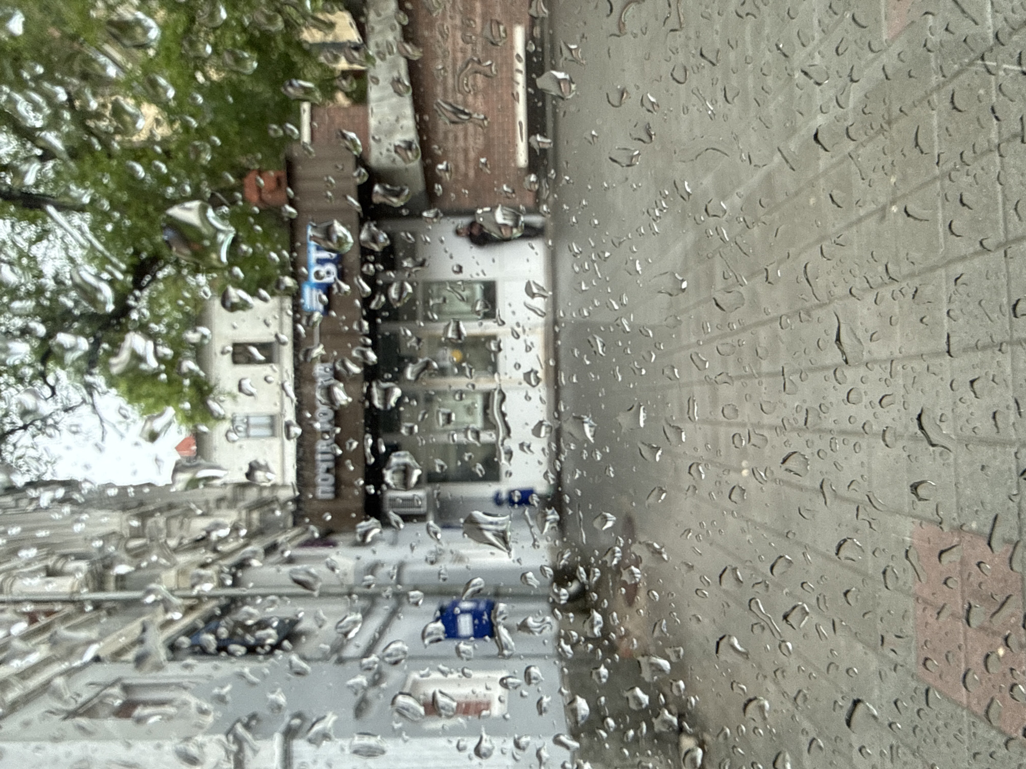 An urban street scene on a rainy day is captured through a window heavily streaked with numerous, glistening raindrops, blurring the background. The image, oriented vertically, shows an outdoor view under natural daylight.
In the foreground, the transparent surface is almost entirely covered by a myriad of irregularly shaped water droplets, creating a soft, distorted filter over the landscape beyond.
Beyond the wet glass, the background reveals an urban environment. To the left, a vibrant green tree with dense foliage partly obscures a light-colored building. A street or paved area extends across the mid-ground, paralleling a building with a dark, possibly brown or reddish-brown, facade. Further to the right, another building or wall with a gray, textured surface is visible.
Among the objects, several blue items, potentially bins or utility boxes, are situated at street level. Metallic structures, possibly railings or construction elements, are also faintly discernible. A faint outline of a person, seemingly wearing dark clothing, stands near the dark-facaded building.
Visible text on the dark building's facade spells out "ПОЧТА" (Pochta) and "РОССИИ" (Rossii), indicative of "Russian Post." A blue sign featuring "B1" or "BI" is also present nearby. The abundant raindrops confirm rainy weather during the daytime. The "Russian Post" signage strongly places the location in Russia, specifically Vladivostok as per the provided context. The overall impression is one of quiet observation from indoors during a rainfall.