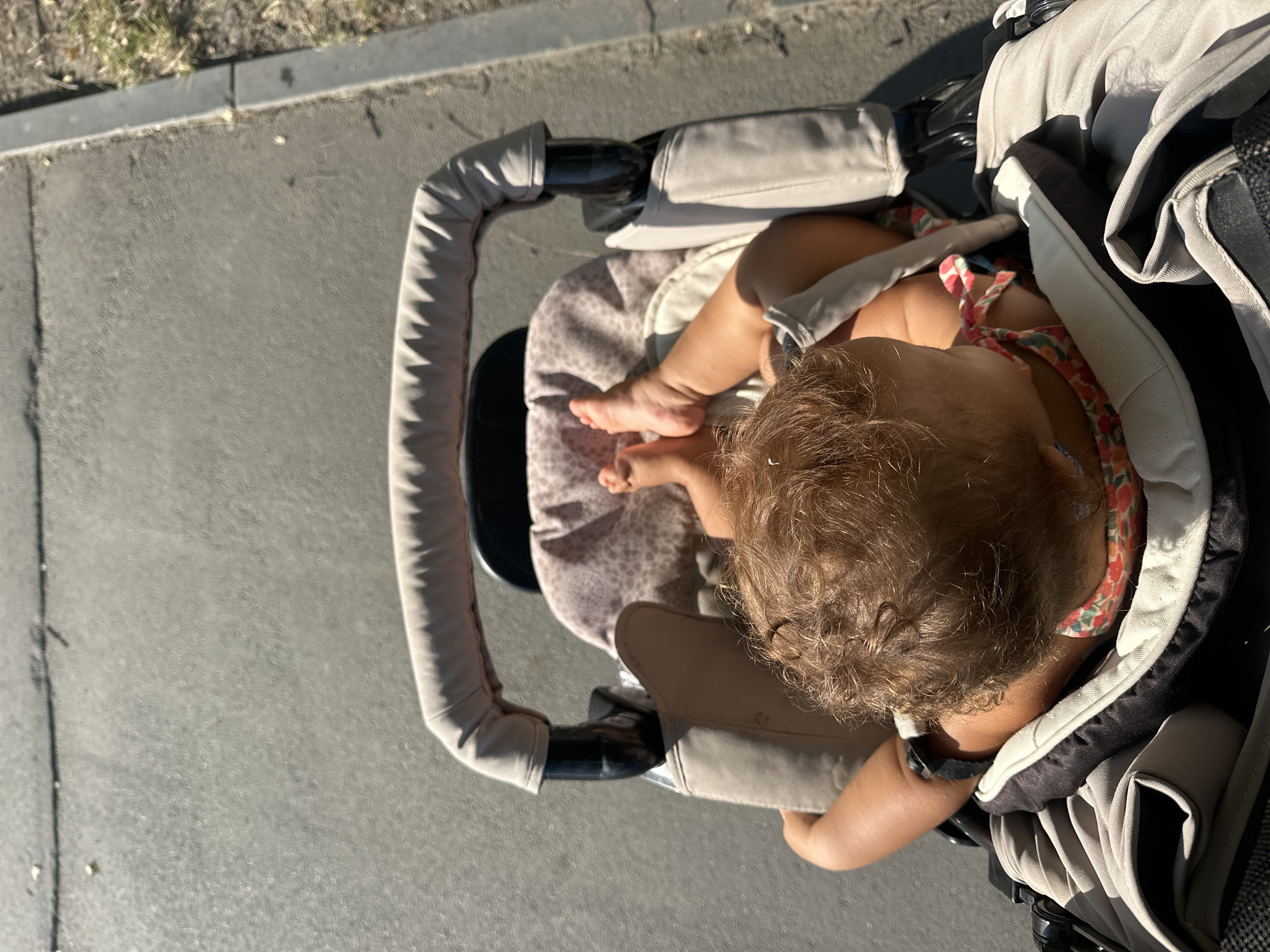 An overhead shot captures a baby in a stroller, viewed from the perspective of the person pushing it. The baby's curly brown hair is prominent, and their small feet are visible in the air. They are wearing a floral-patterned outfit. The stroller itself is light beige with black accents and a padded handle. The background consists of a paved surface, likely a sidewalk or path, with a slight incline and a curb visible on the right side, adjacent to dry grass and dirt. The lighting suggests a sunny day. The image does not contain any specific landmarks or features to determine its exact location within Petrozavodsk, Russia.