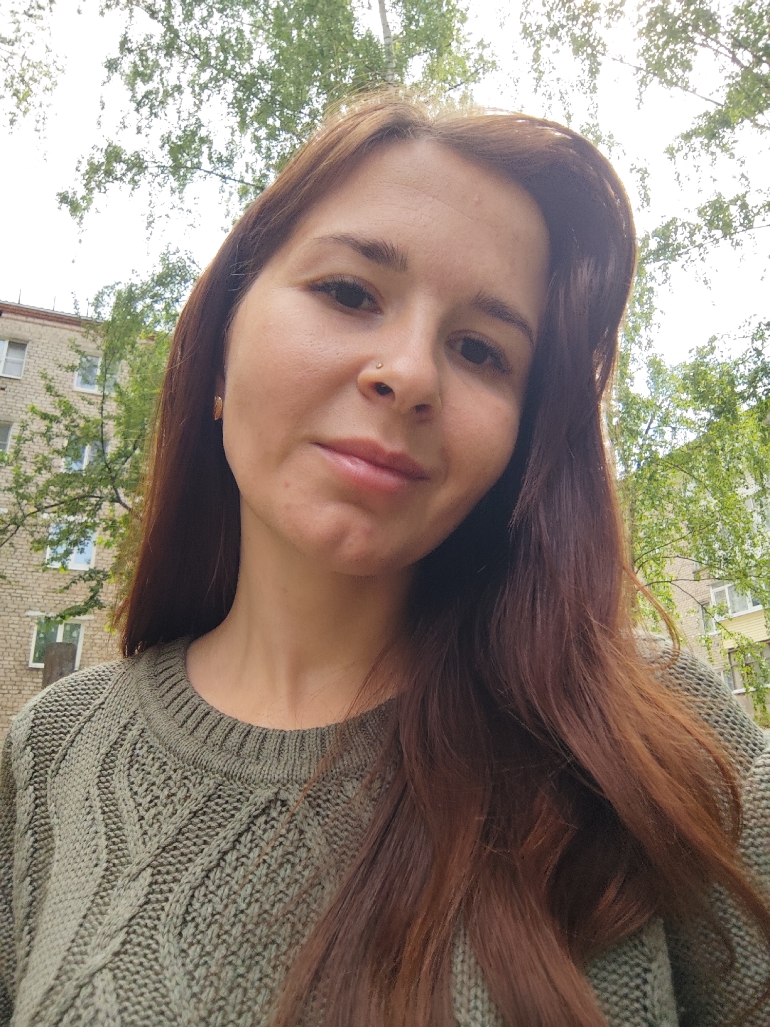 A young woman with long, auburn hair, wearing a green knit sweater, is standing outside. She is looking at the camera with a slight smile. A building is visible in the background, and trees with green leaves surround the woman and the building. The building is likely located in the city of Chaikovsky, Russia.